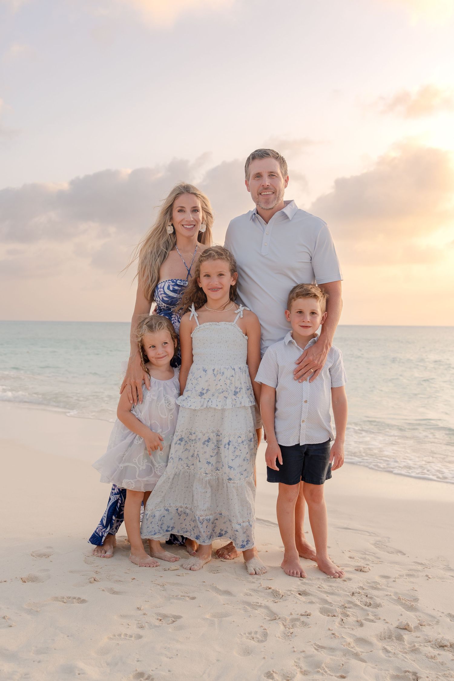 Family of five posing together on a sandy beach at sunset, parents with three children in coordinating white outfits.