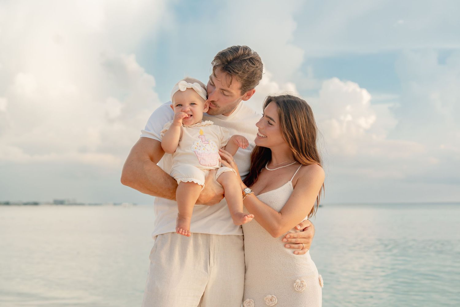 A happy young family with a baby poses on a serene beach with calm blue water and soft clouds behind them.