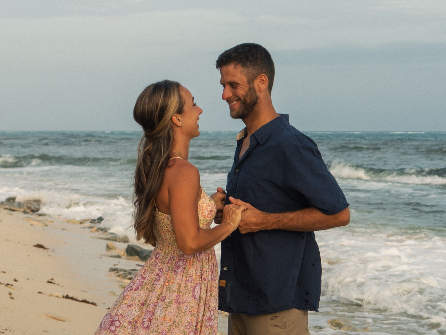 Happy couple holding hands and smiling at each other on a beautiful sandy beach with ocean waves.