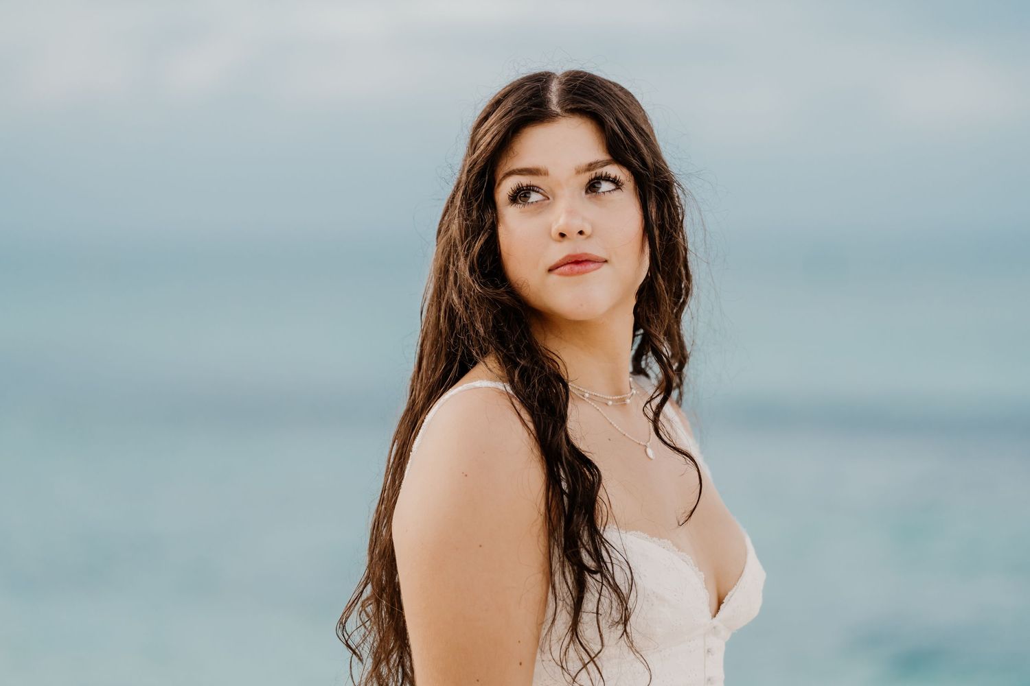 A person in a white dress stands on a beach with ocean waves and blue sky visible.