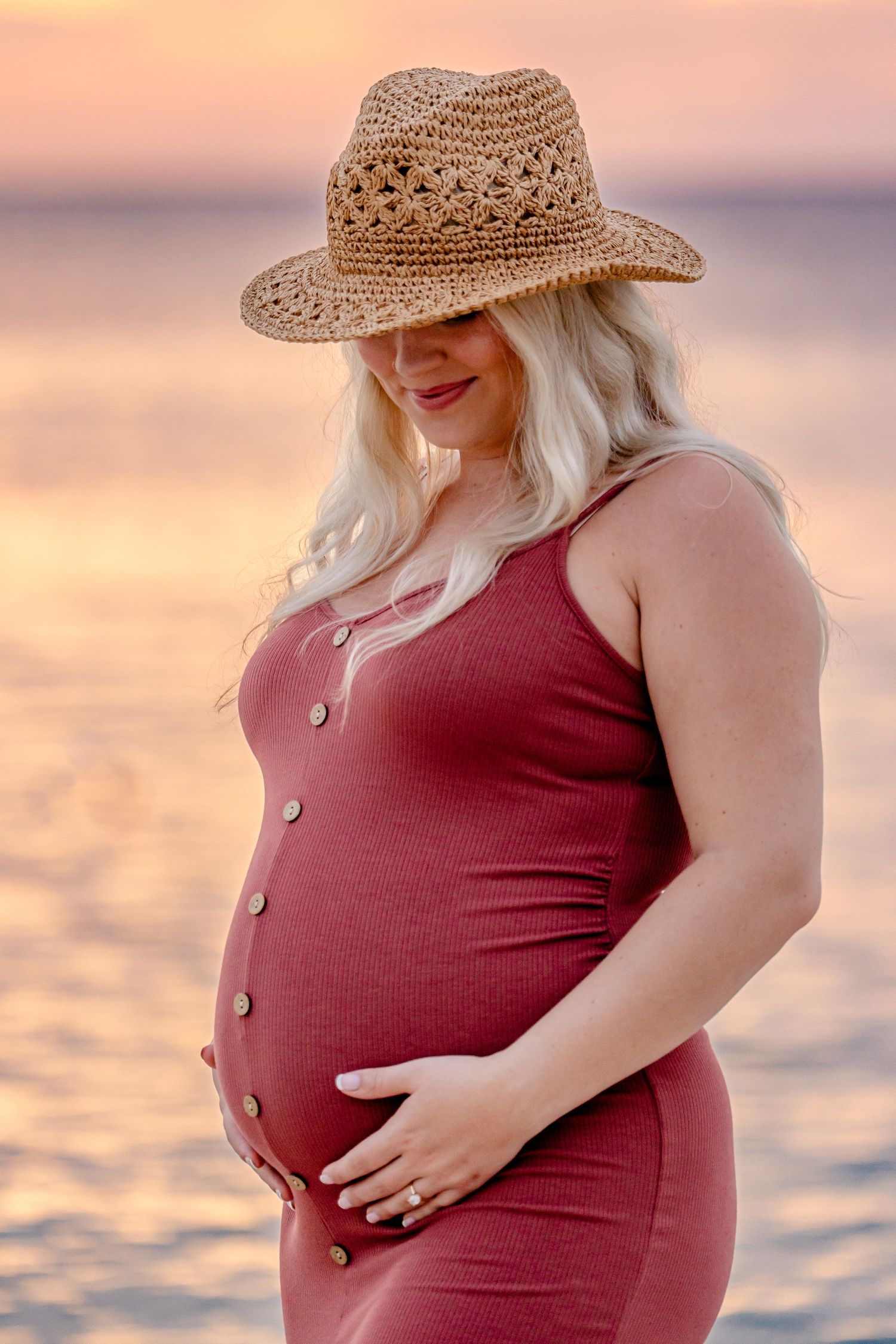 Maternity photo at sunset wearing red dress and straw hat by the ocean.