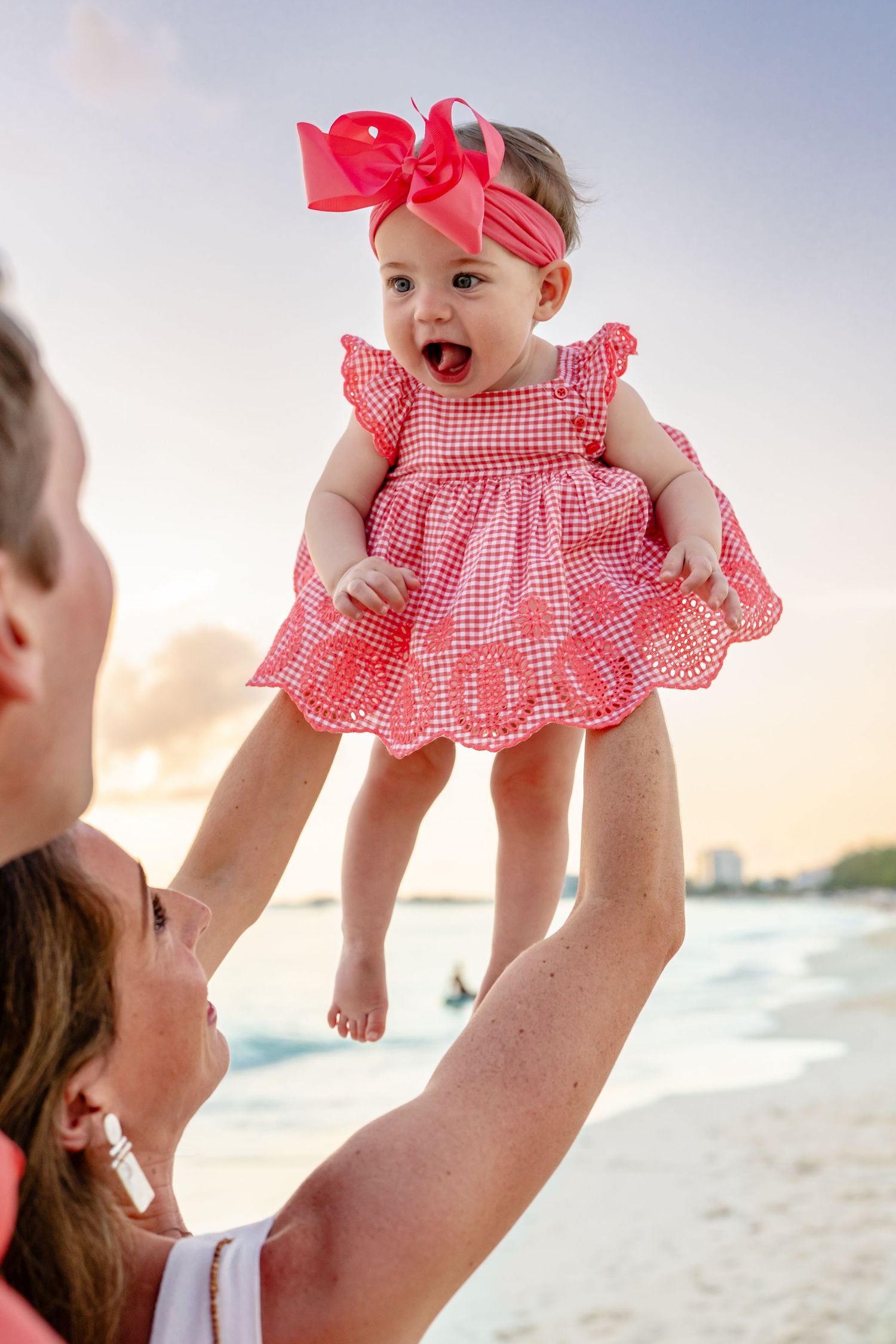 Baby in red gingham dress being lifted joyfully in the air at sunset on the beach.