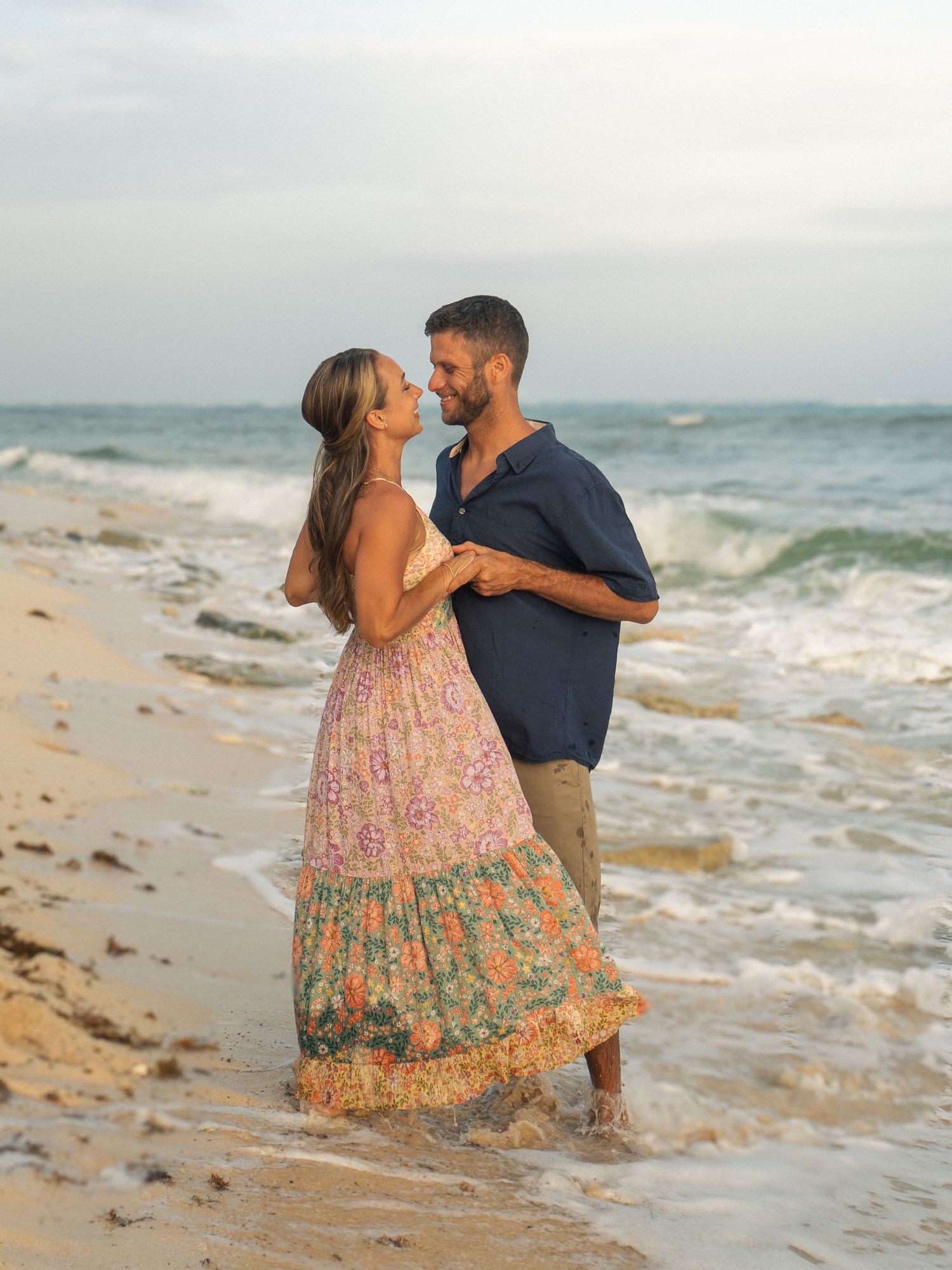 Couple dancing romantically on a sandy beach in the ocean waves at dusk, woman in floral maxi dress.