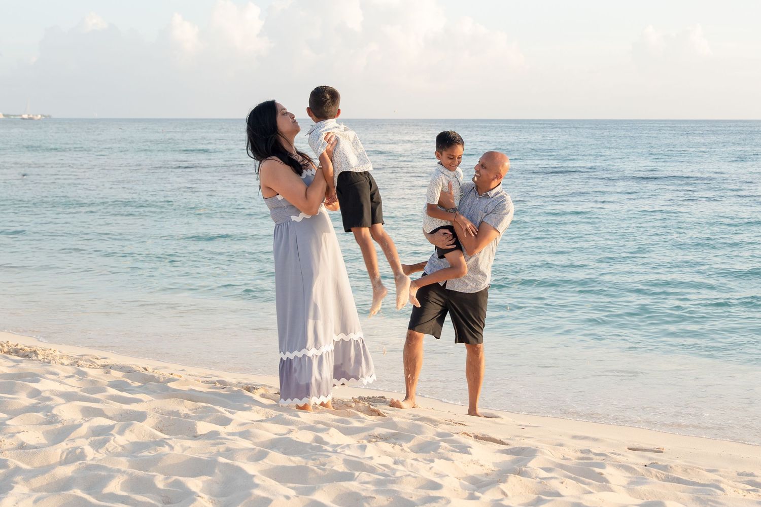 Family of four enjoying a sunny beach day, with parents playfully lifting their children on white sand by calm turquoise waters.