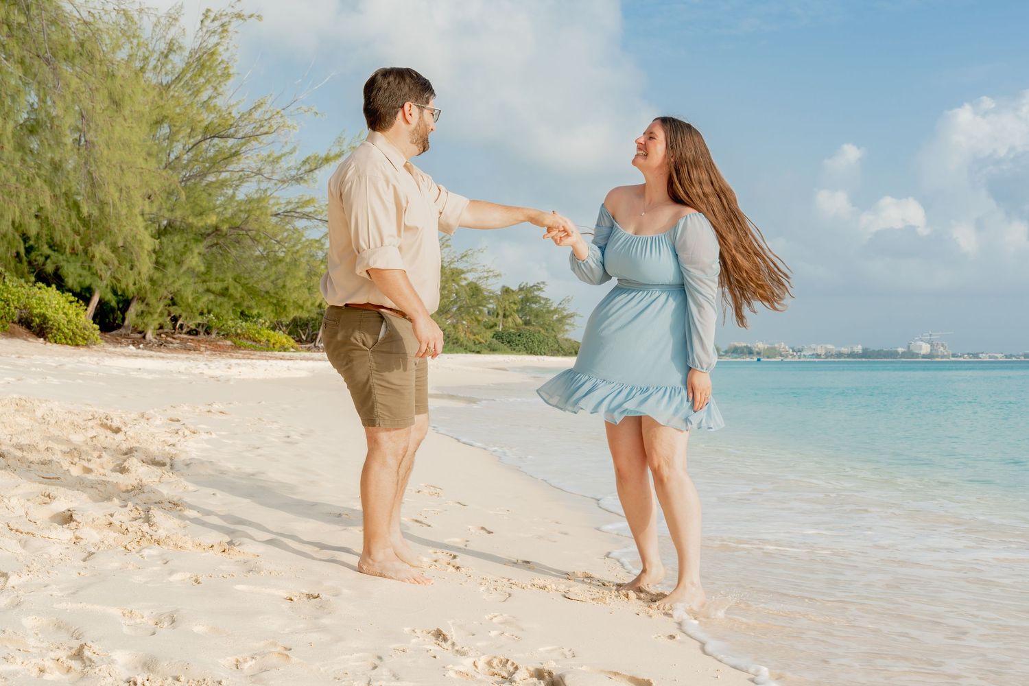 A couple holds hands and dances barefoot on a white sandy beach with turquoise water and blue sky.