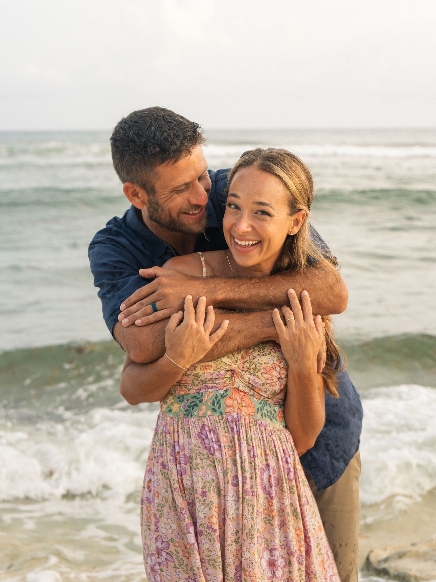 Happy couple embracing on a beach with ocean waves, woman in floral dress laughing joyfully.