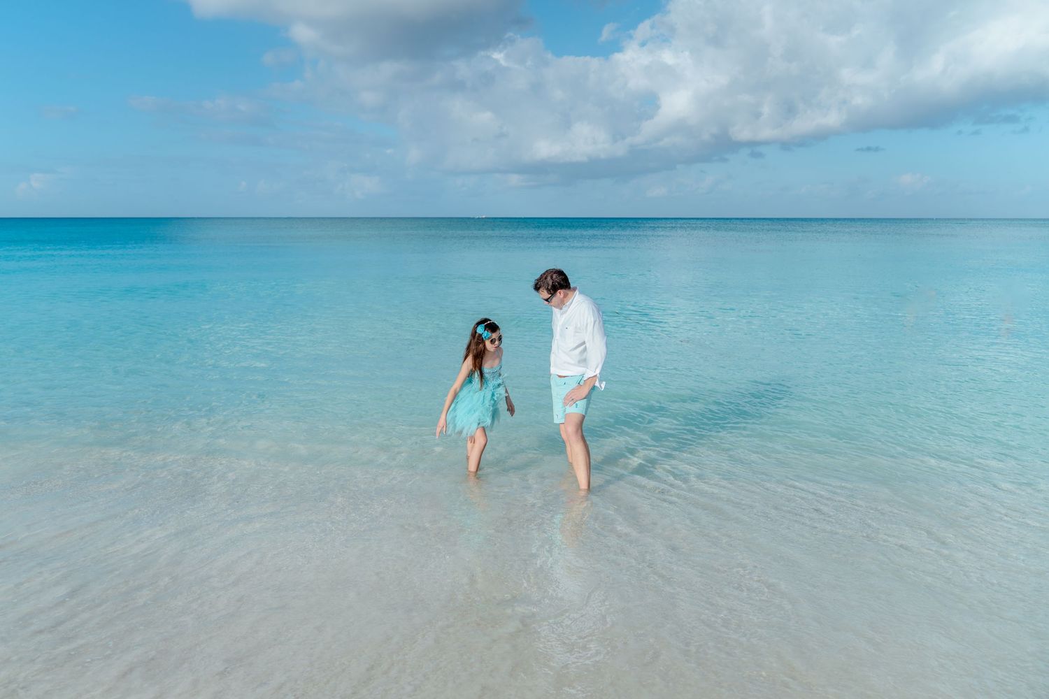 People walking in crystal clear turquoise waters along a pristine beach with white puffy clouds in the sky.