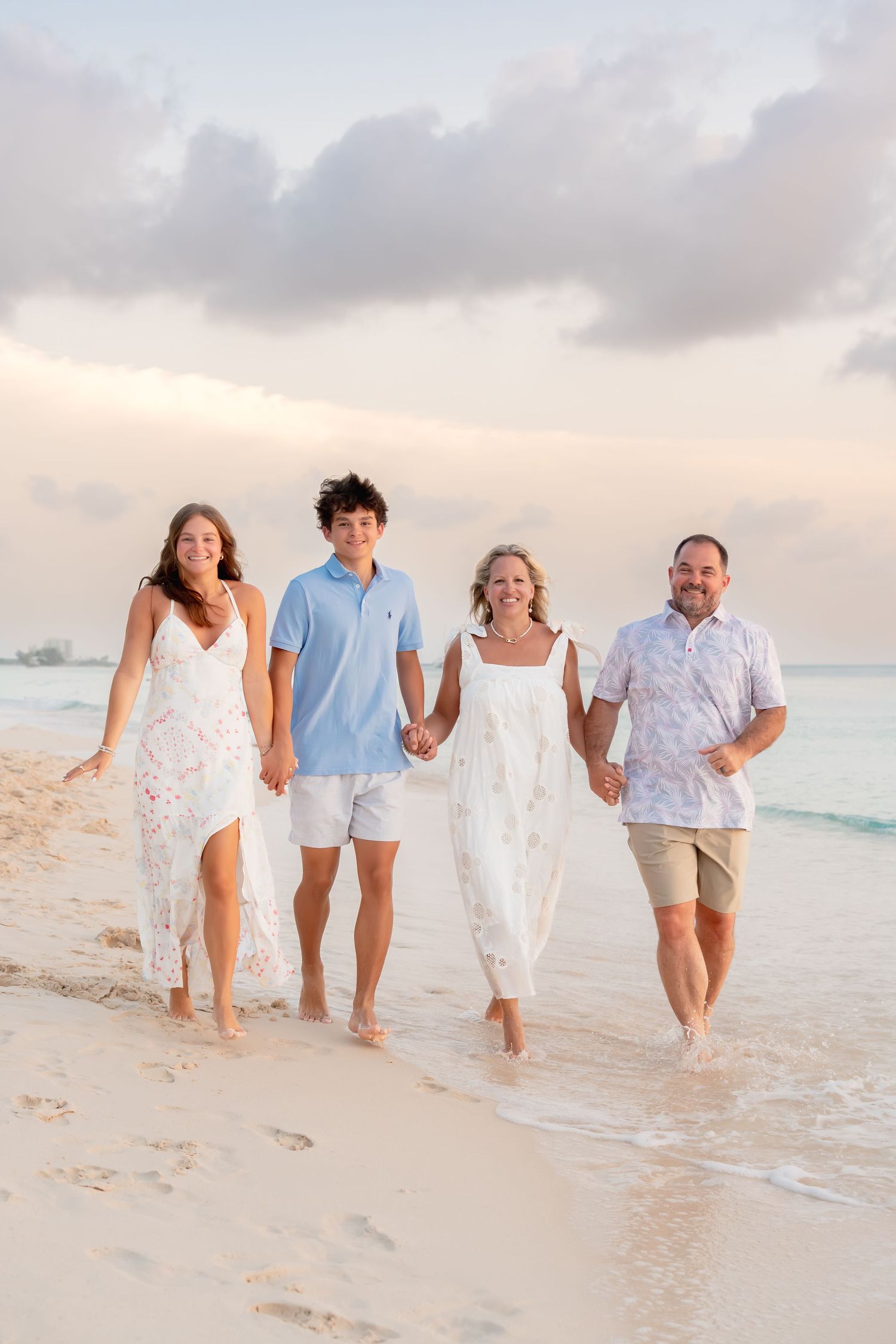 Family of four walking on a sandy beach at sunset, wearing white and pastel outfits, smiling and holding hands.