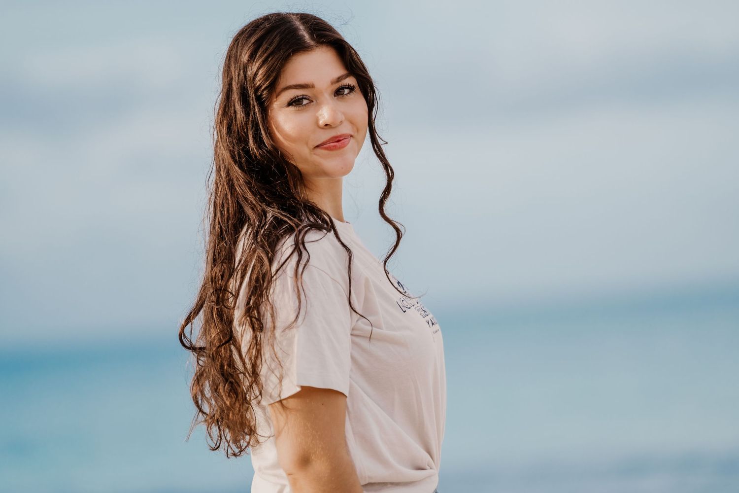 Young woman in casual attire poses against a cloudy beach backdrop.