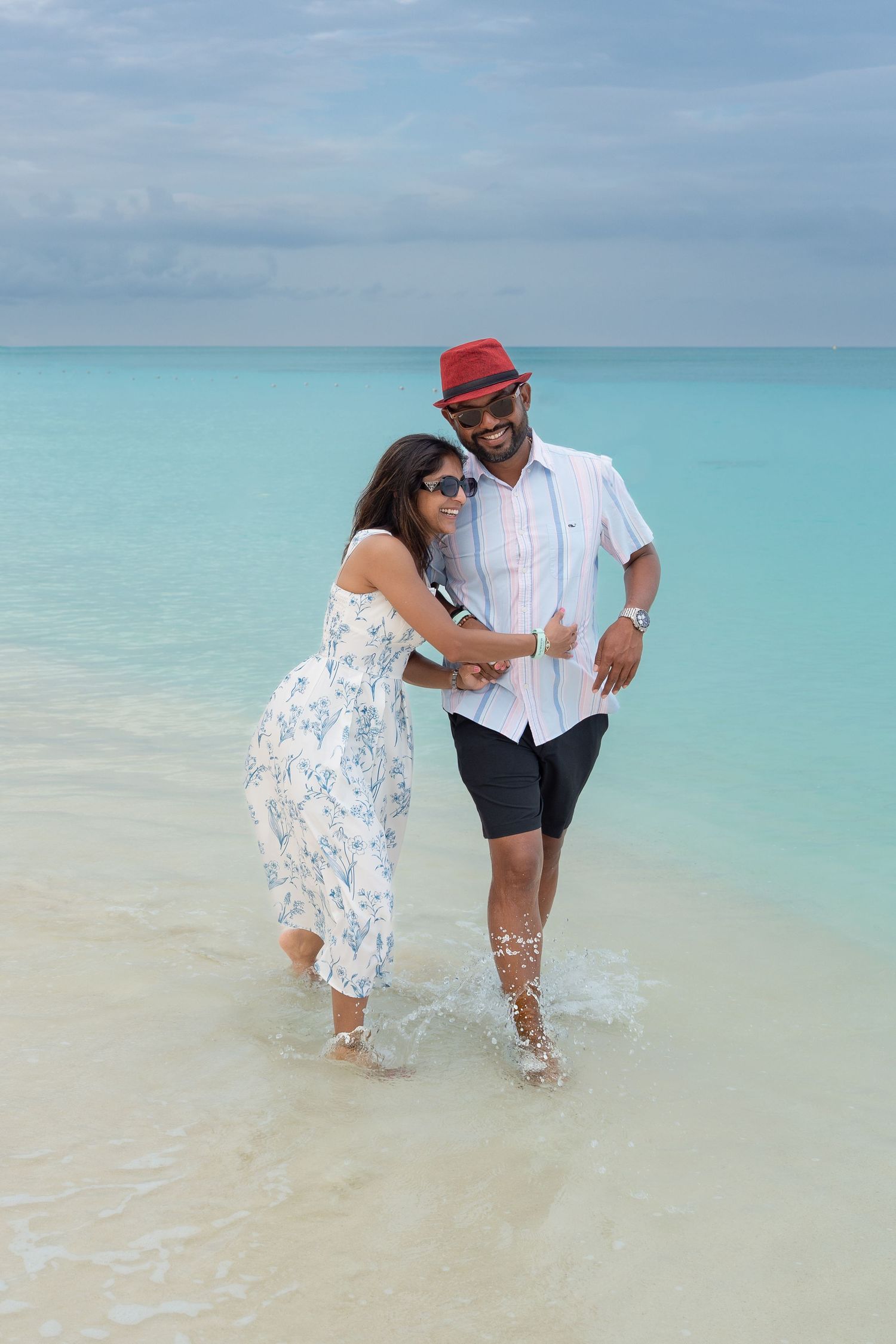 Happy couple in sunglasses wading in turquoise ocean water on a tropical beach, woman in floral dress, man in red hat.
