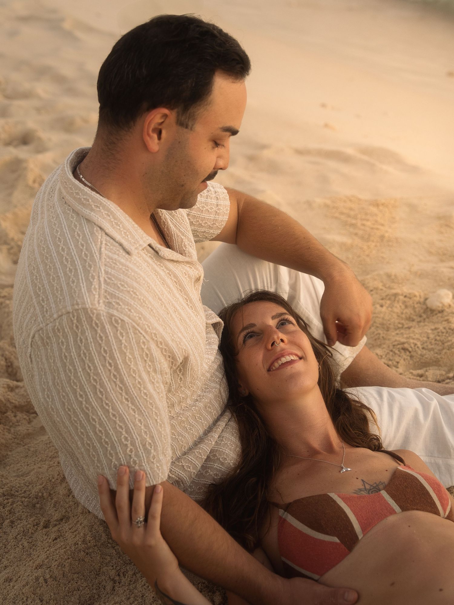 Couple relaxing on sandy beach at sunset, woman lying down smiling up at her partner.