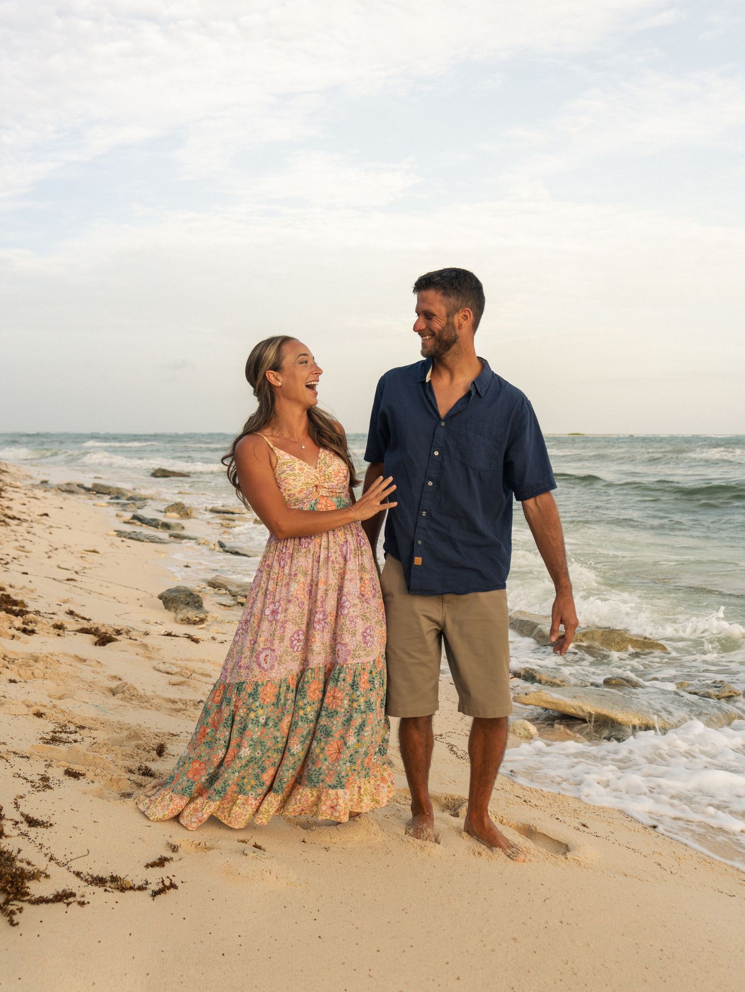 Couple laughing and walking barefoot on a sandy beach, woman in floral maxi dress, man in navy shirt and khaki shorts.
