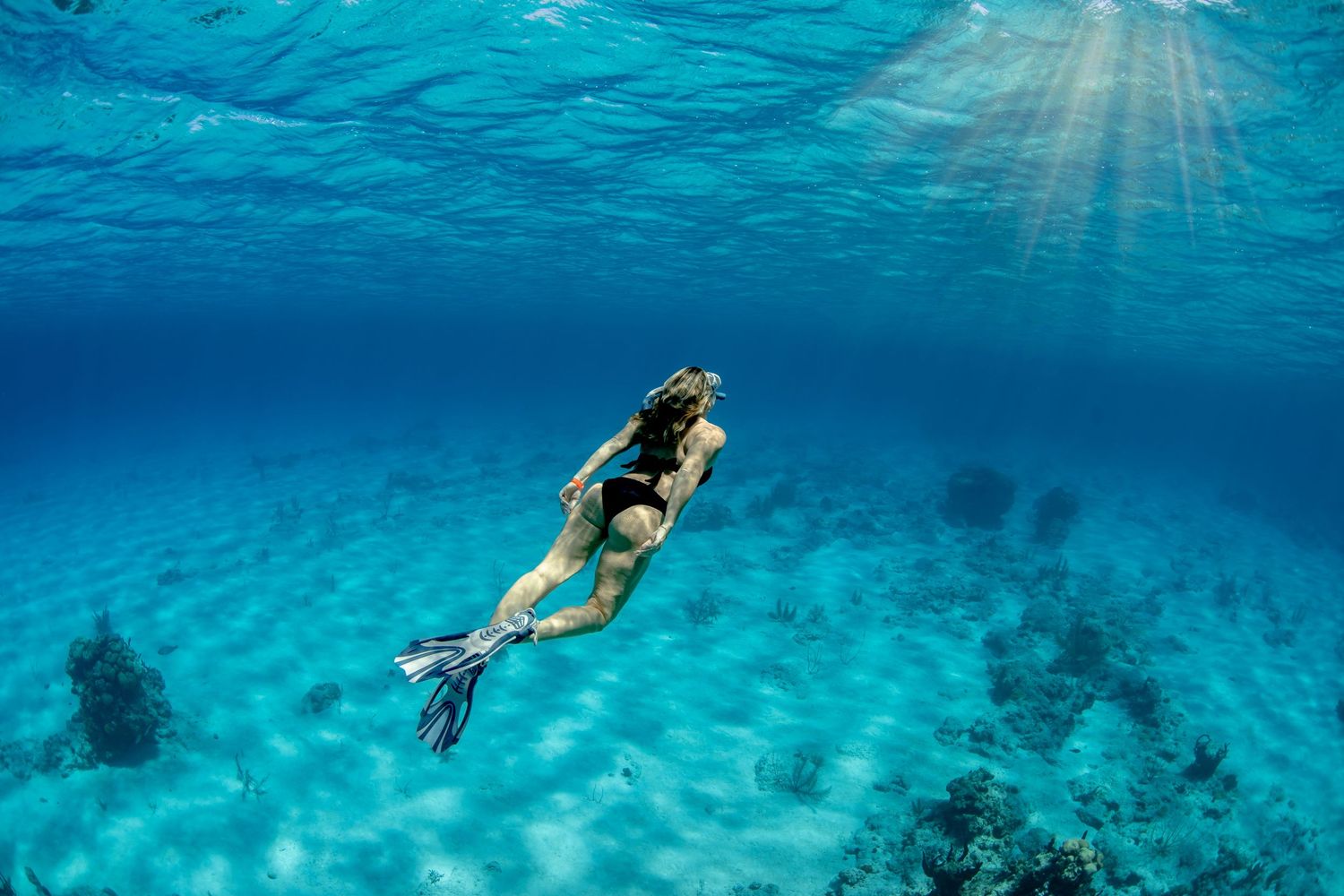 Snorkeler swimming in clear turquoise waters above coral reef with sunbeams streaming down.