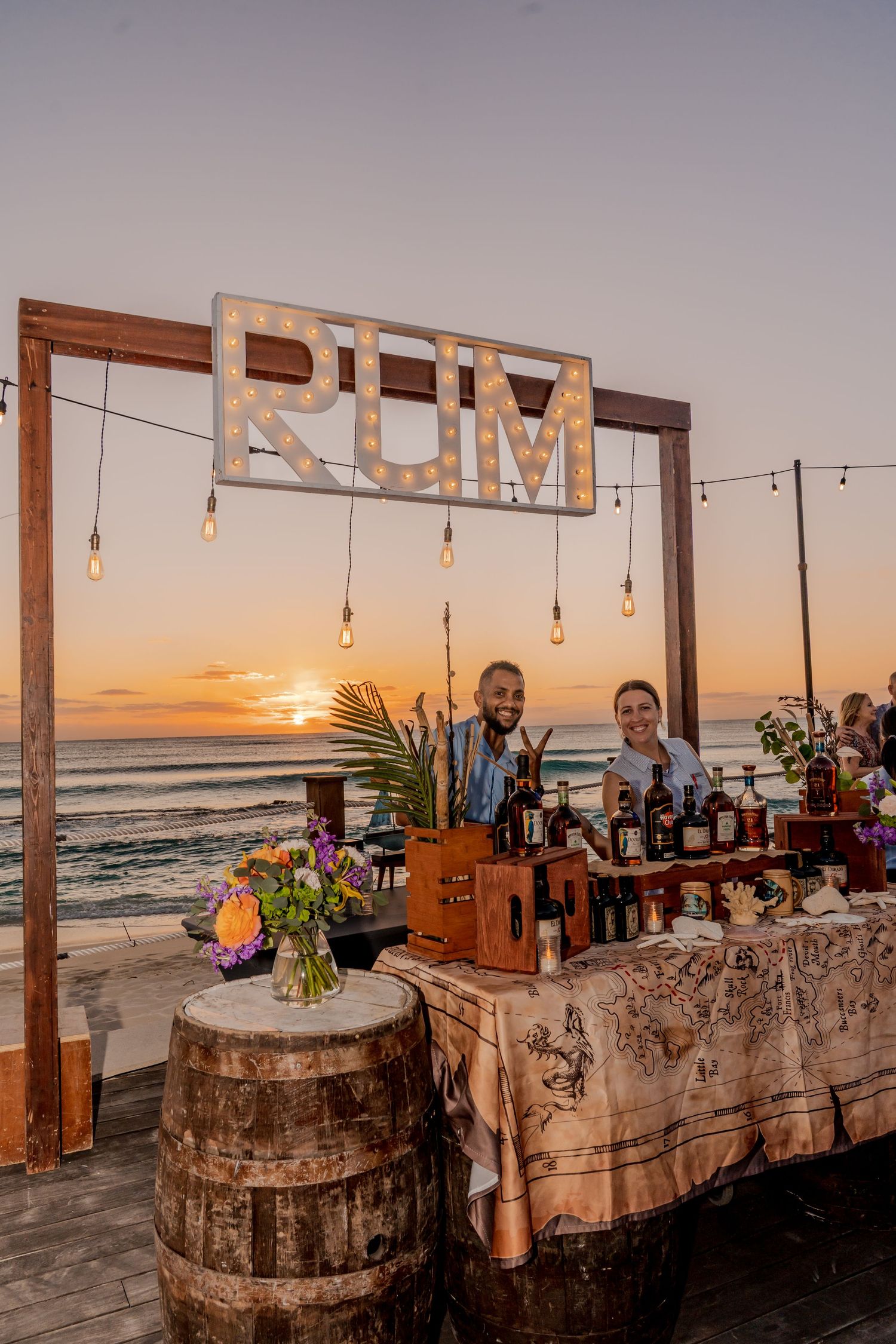 Rustic beach bar setup with wooden signs and string lights at sunset overlooking the ocean.