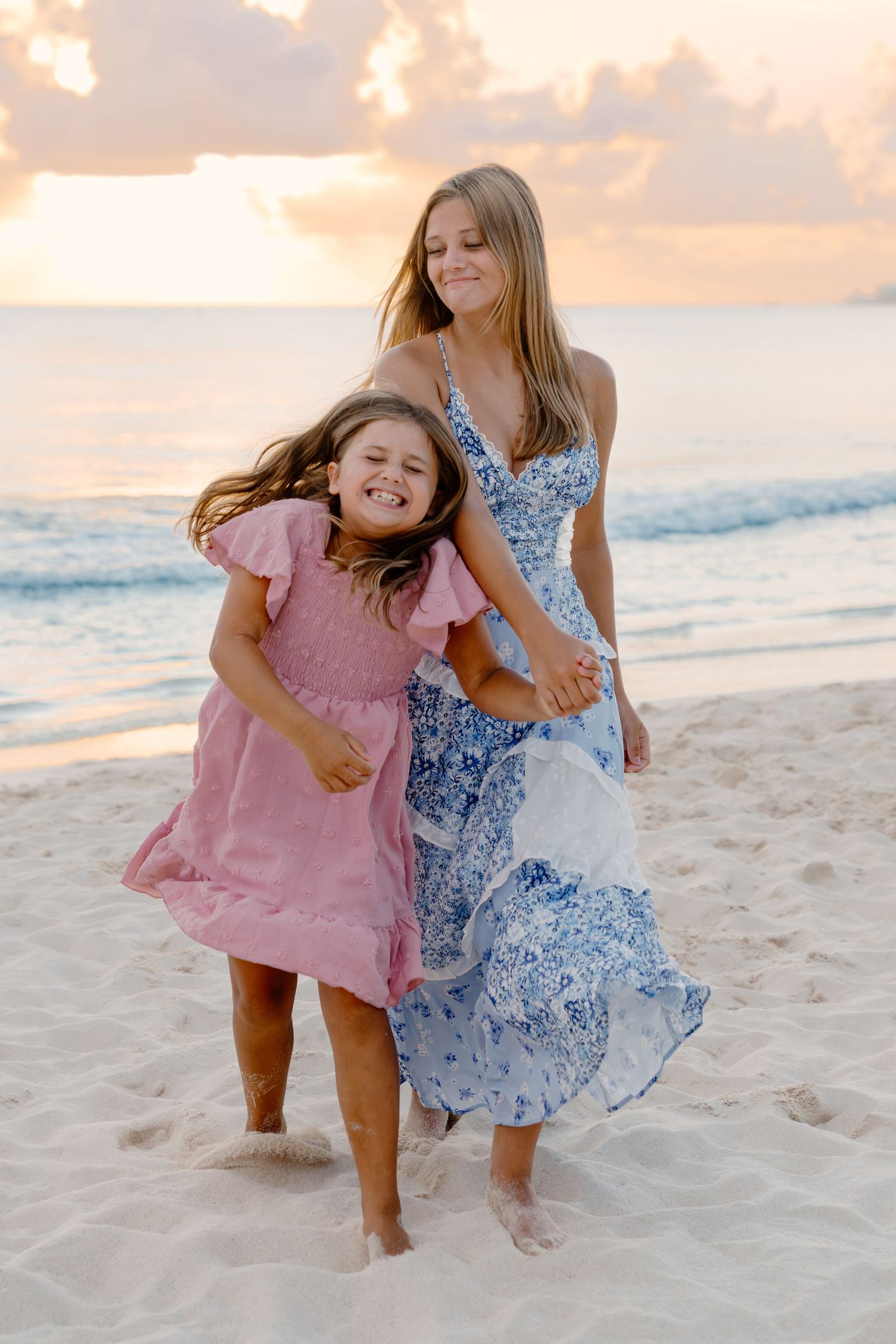 Two people in flowy dresses play and laugh together on a sandy beach at sunset.