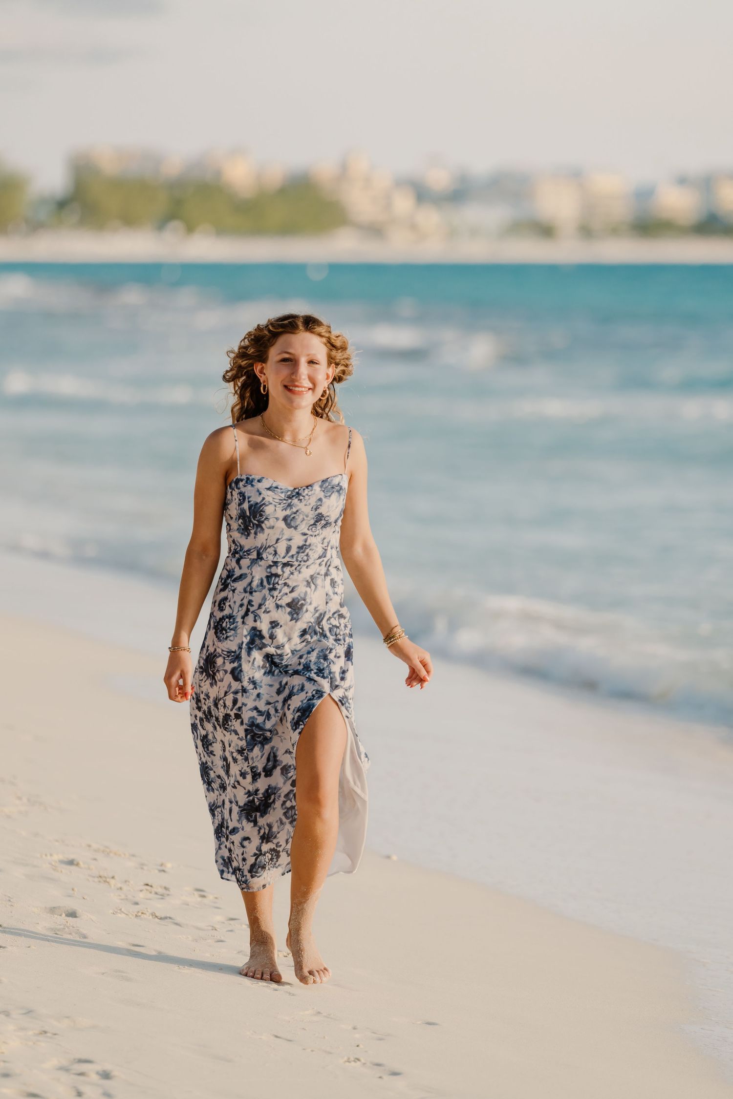 Person in floral dress running playfully along white sandy beach with ocean waves.