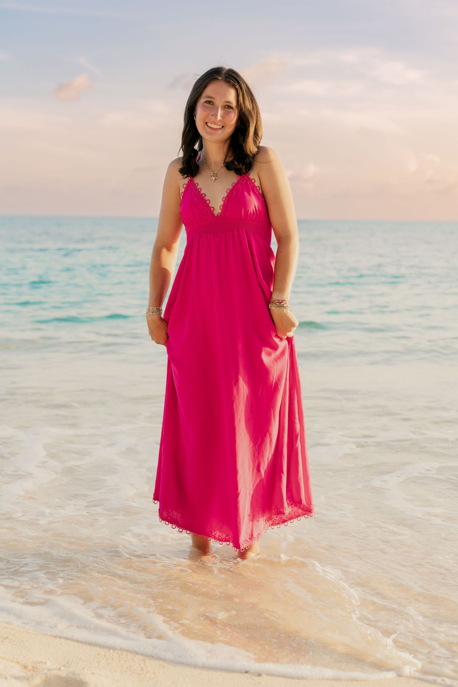 A figure in a flowing hot pink dress stands at the edge of turquoise ocean waters during sunset.