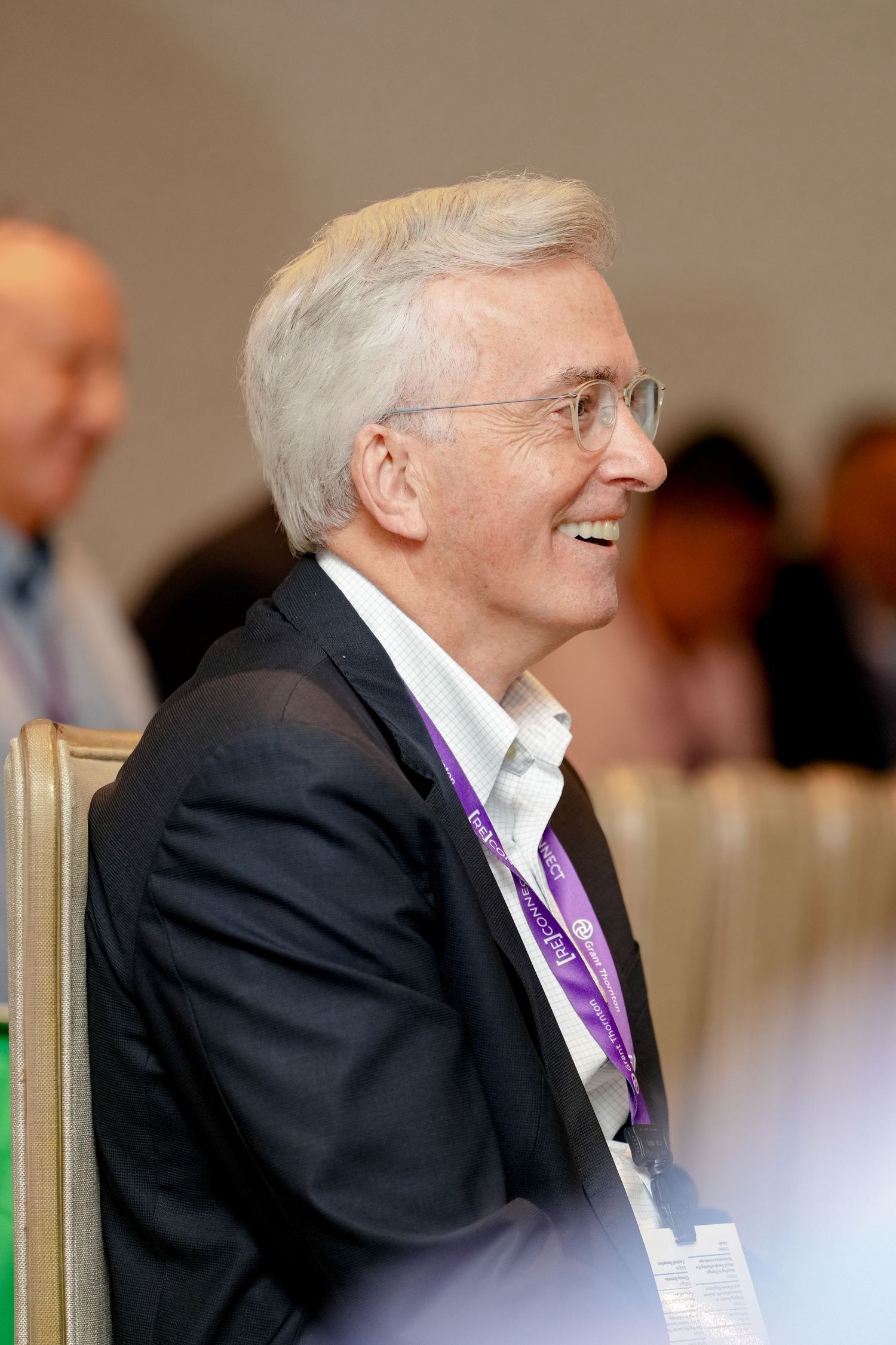 Senior executive in a dark suit and purple lanyard smiling during a business conference.