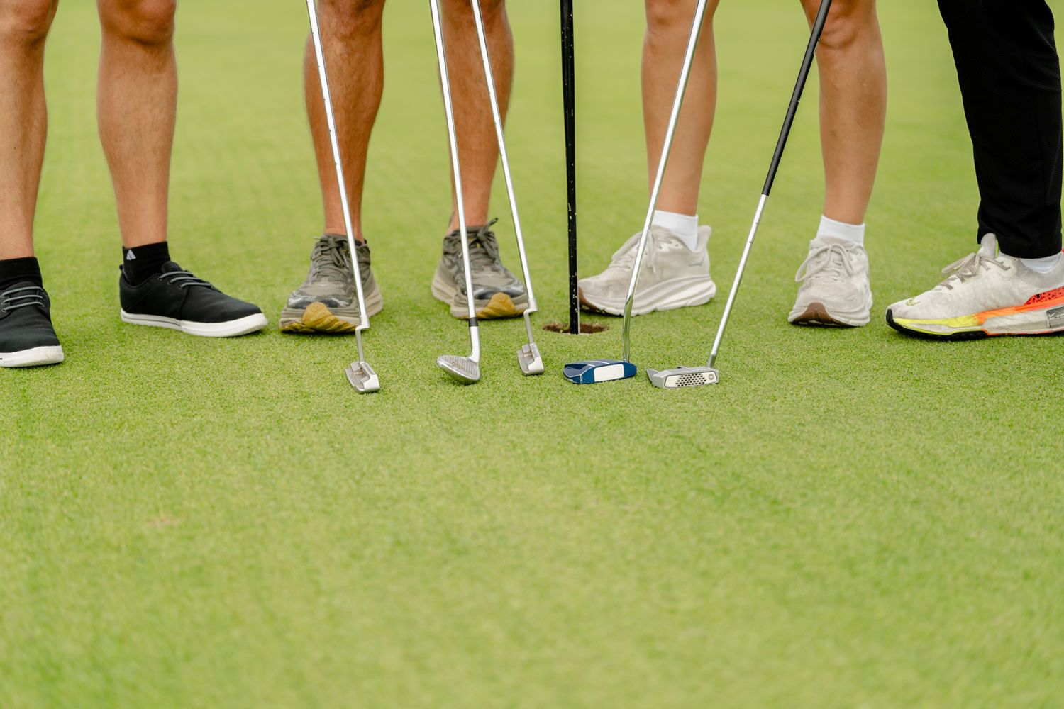 Close-up of golf clubs and shoes on putting green showing multiple players preparing to putt.