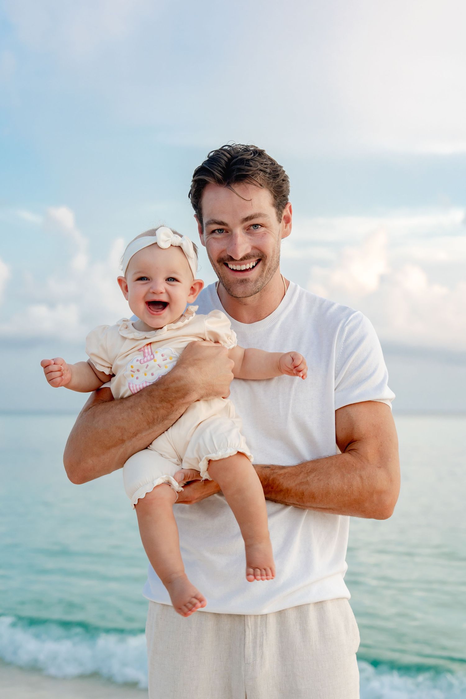 Happy father holding laughing baby girl on a sunny beach with turquoise ocean waves in the background.
