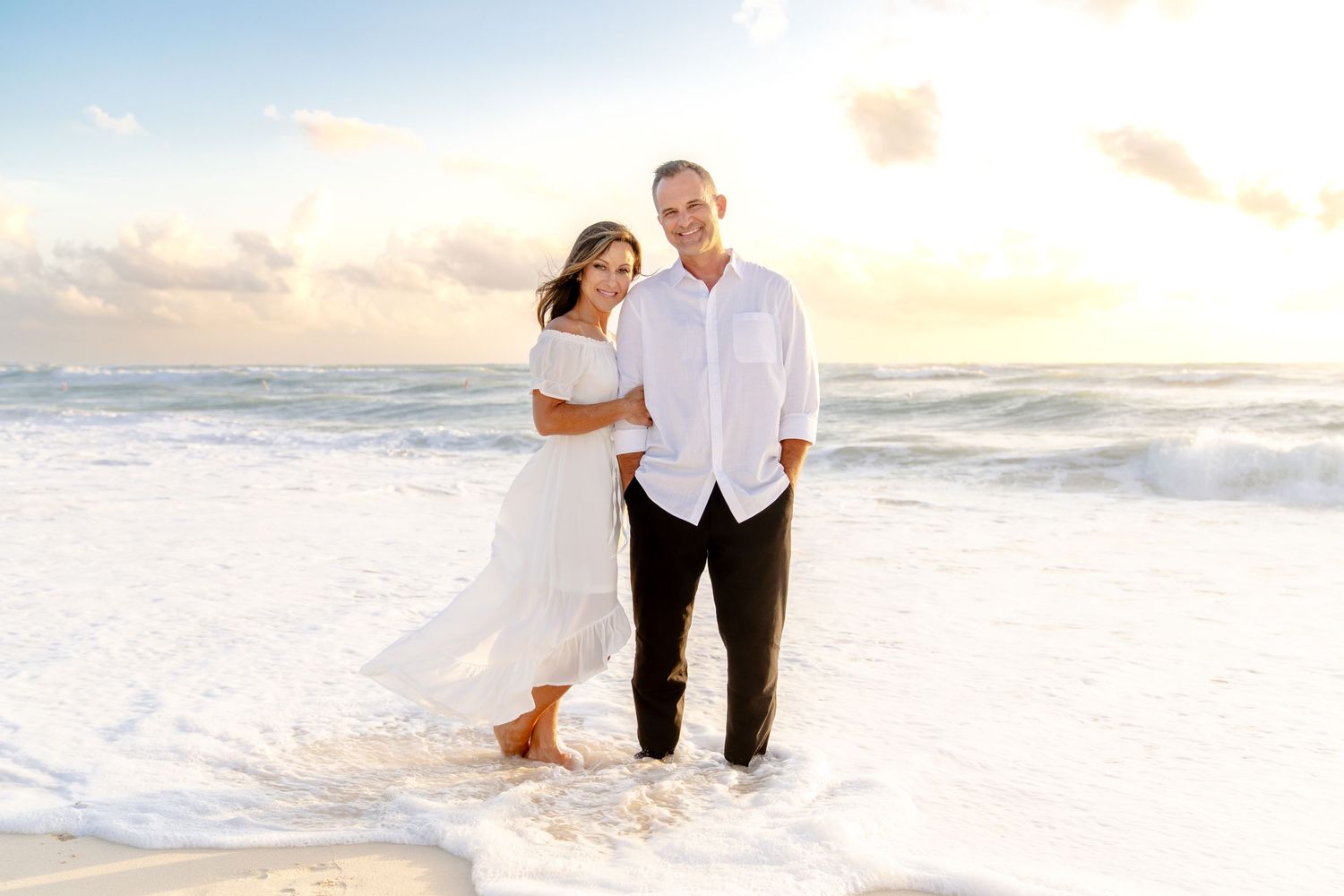 Romantic beach photoshoot at sunset with couple walking along the shoreline in white attire.