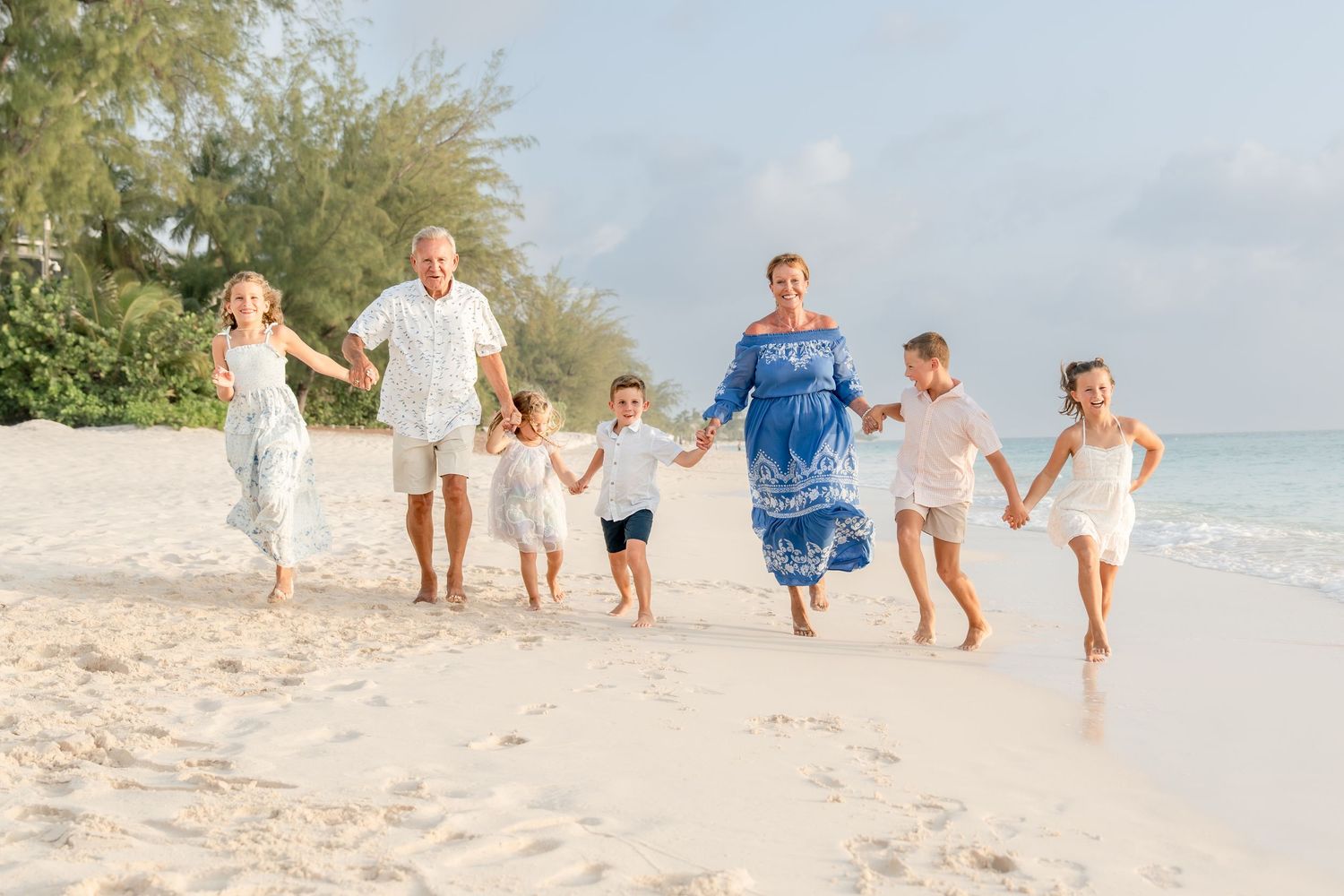 Family of six joyfully running on a sunny white sand beach, wearing white and blue outfits near turquoise ocean waters.
