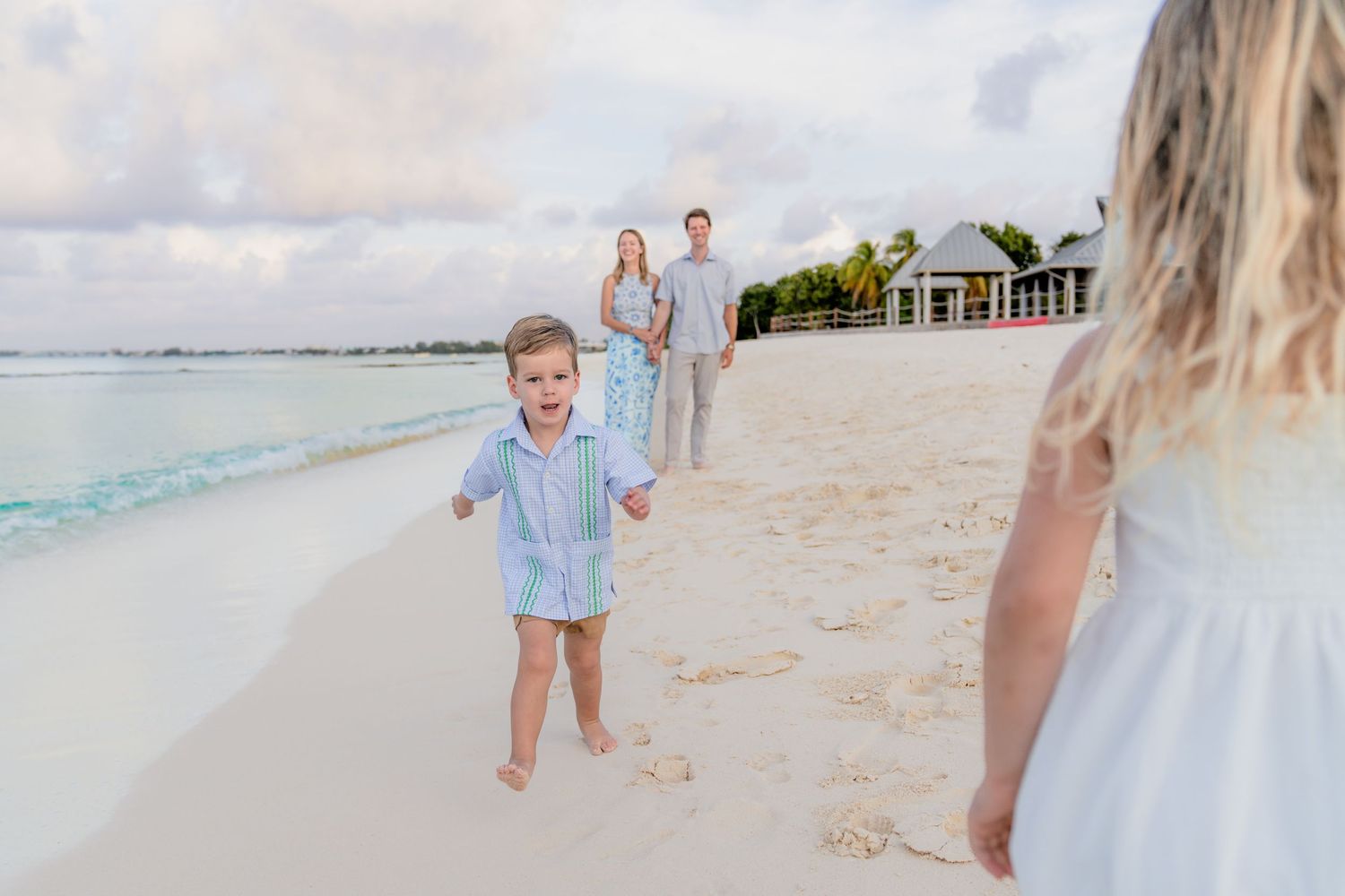 A family walks along a pristine white sand beach at sunset with turquoise waters and a pier in the background.