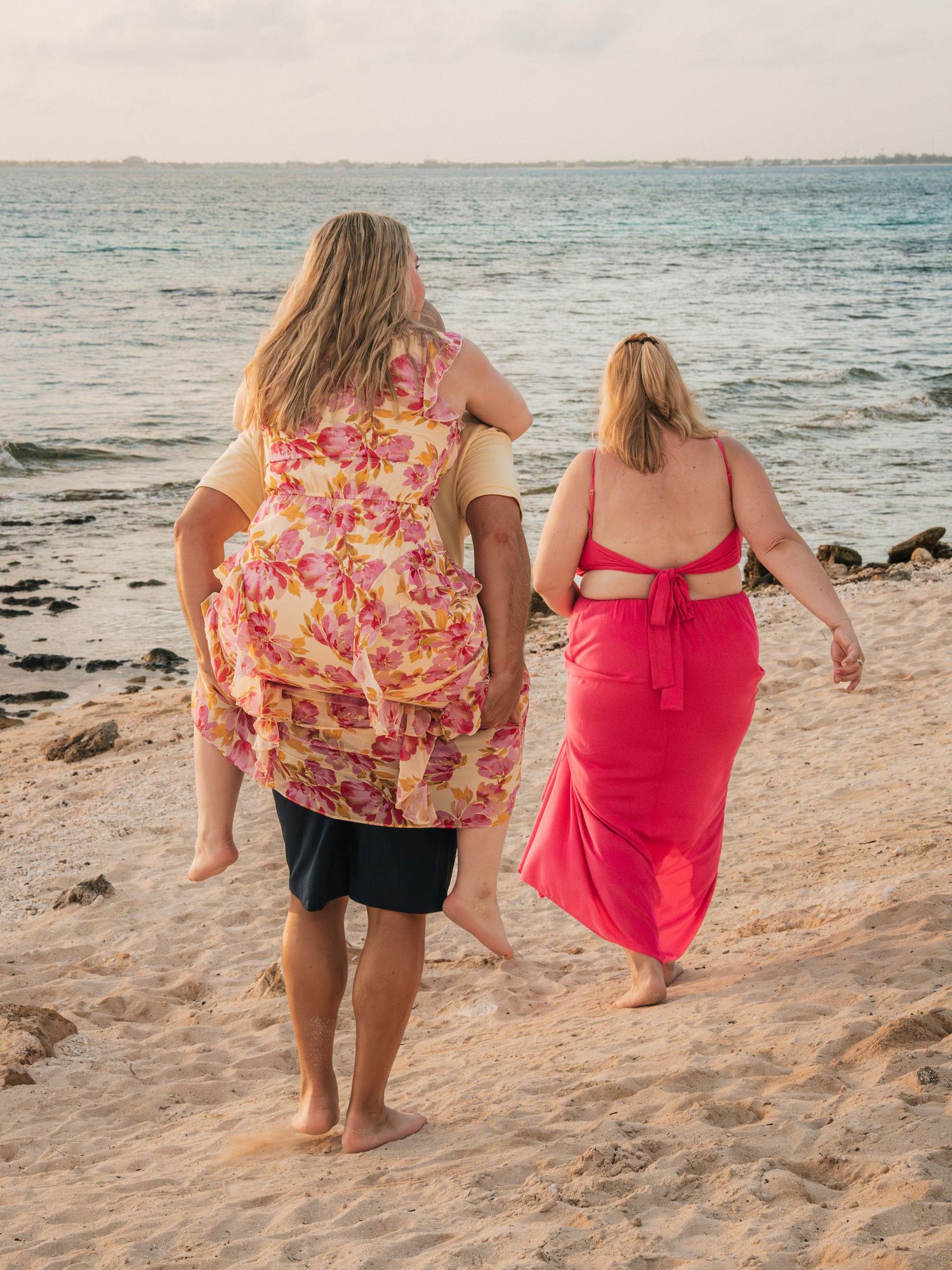 Family walking barefoot on sandy beach, man carrying girl in floral dress, woman in pink maxi dress.