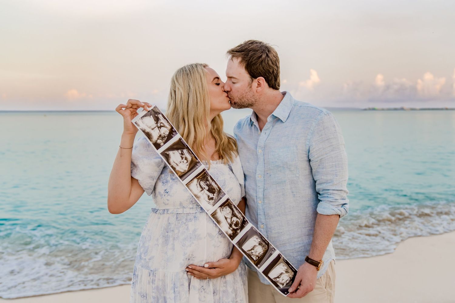 Couple holding ultrasound photos while sharing tender moments on sandy beach.