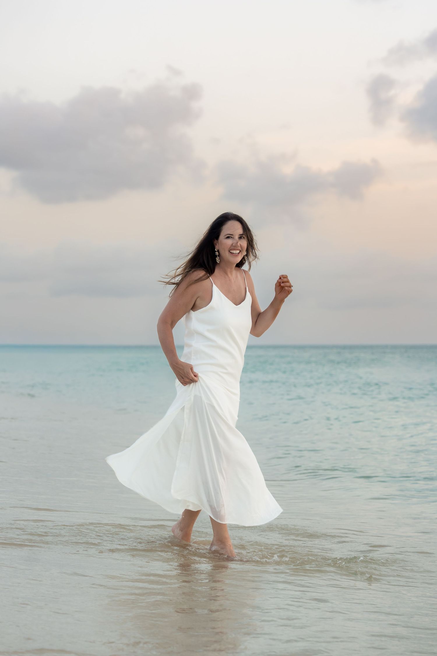 Woman in white flowing dress walking barefoot on a tropical beach at sunset, smiling joyfully.