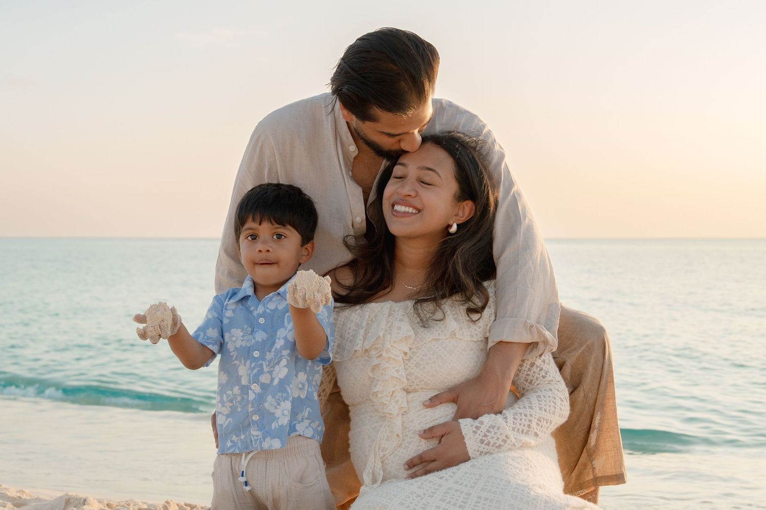 Pregnant woman in white lace dress smiles with husband and toddler son on a beach at sunset.