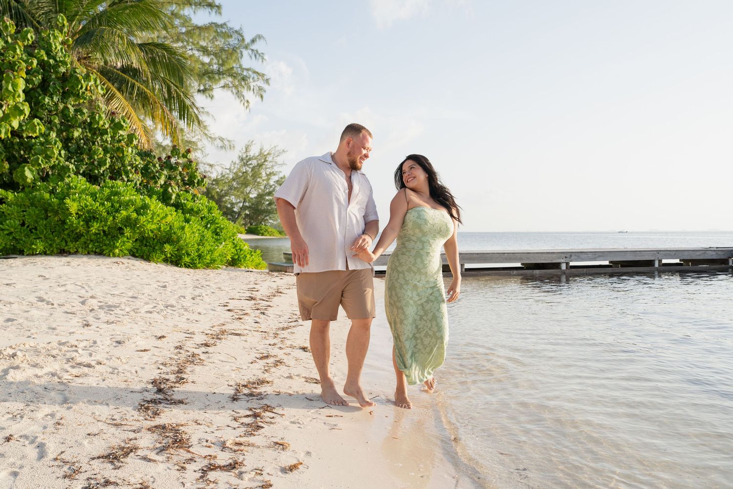 Couple holding hands walking barefoot on a tropical beach with palm trees and clear blue water.