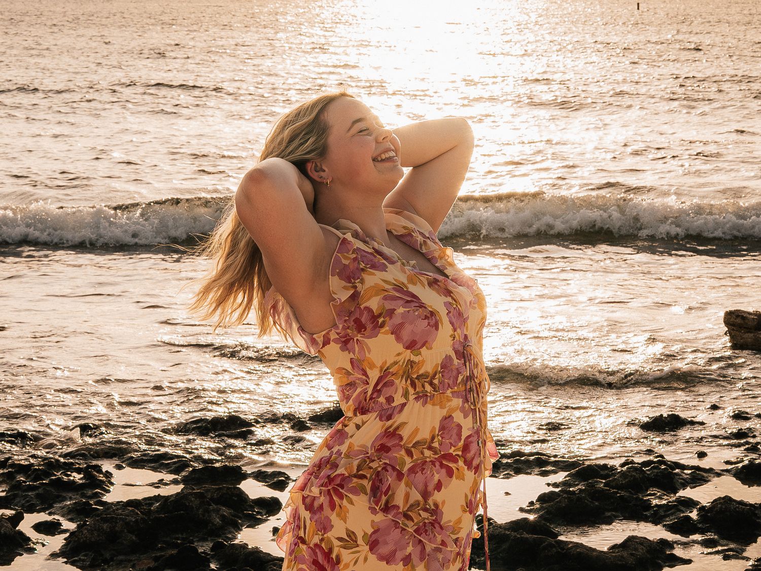 Woman in floral dress laughing on rocky beach at golden sunset, hair blowing in the breeze.