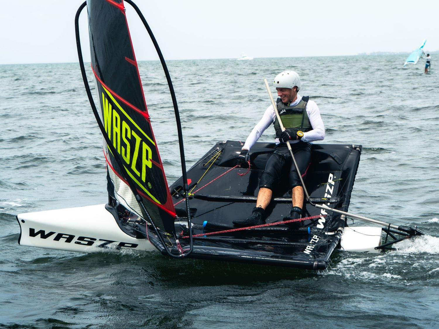 Sailor navigating a WASZP foiling sailboat on choppy water during a race or training session.