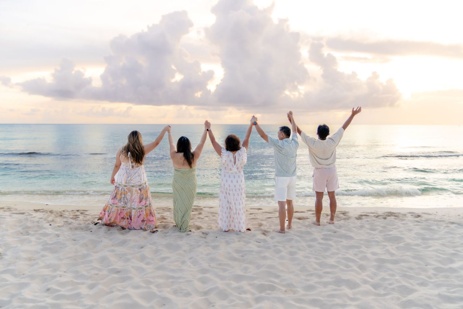 Group celebrating with raised arms on beach at sunset wearing coordinated white and floral outfits.