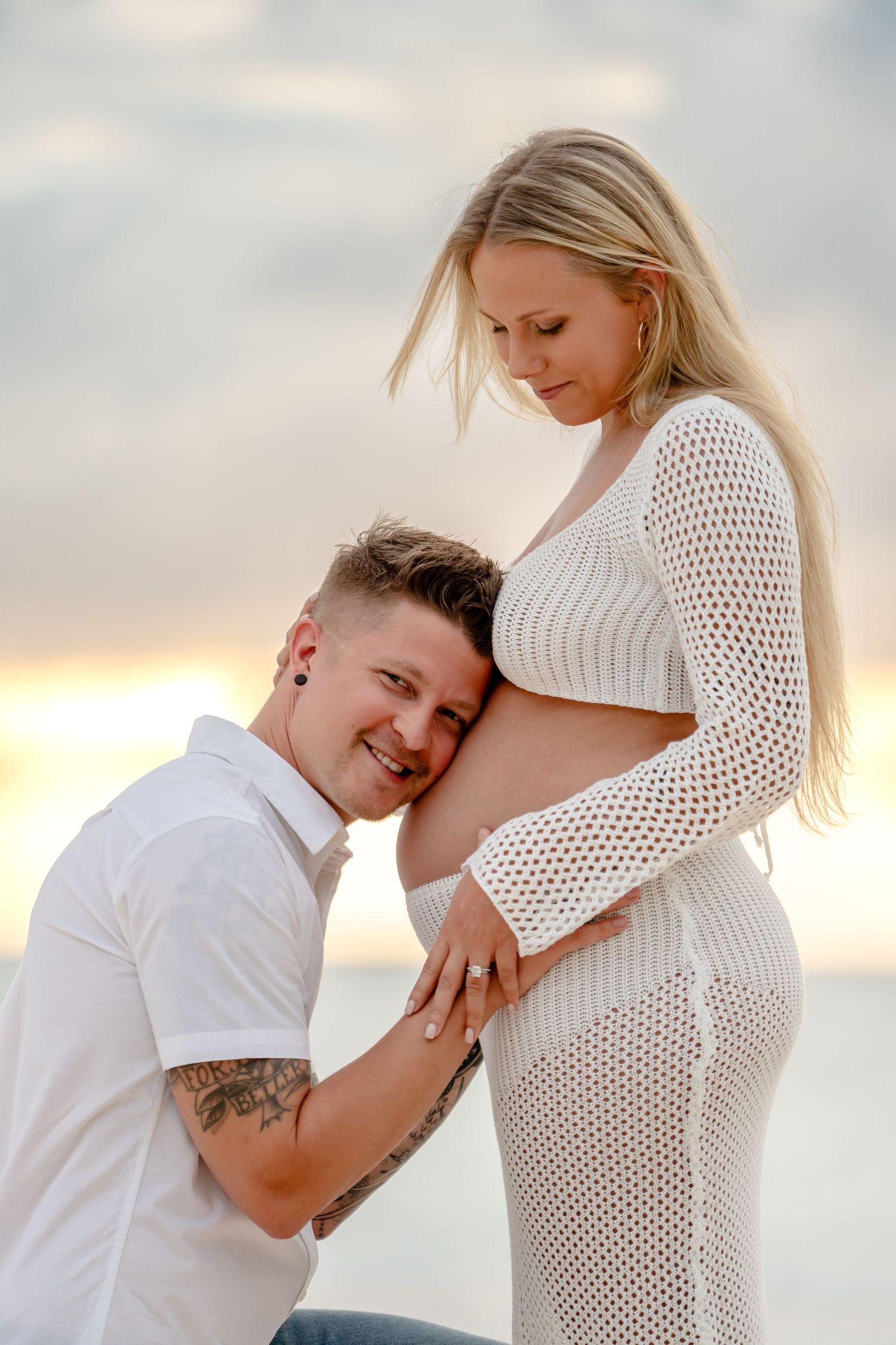 Tender maternity moments captured on beach with couple in white clothing at sunset.