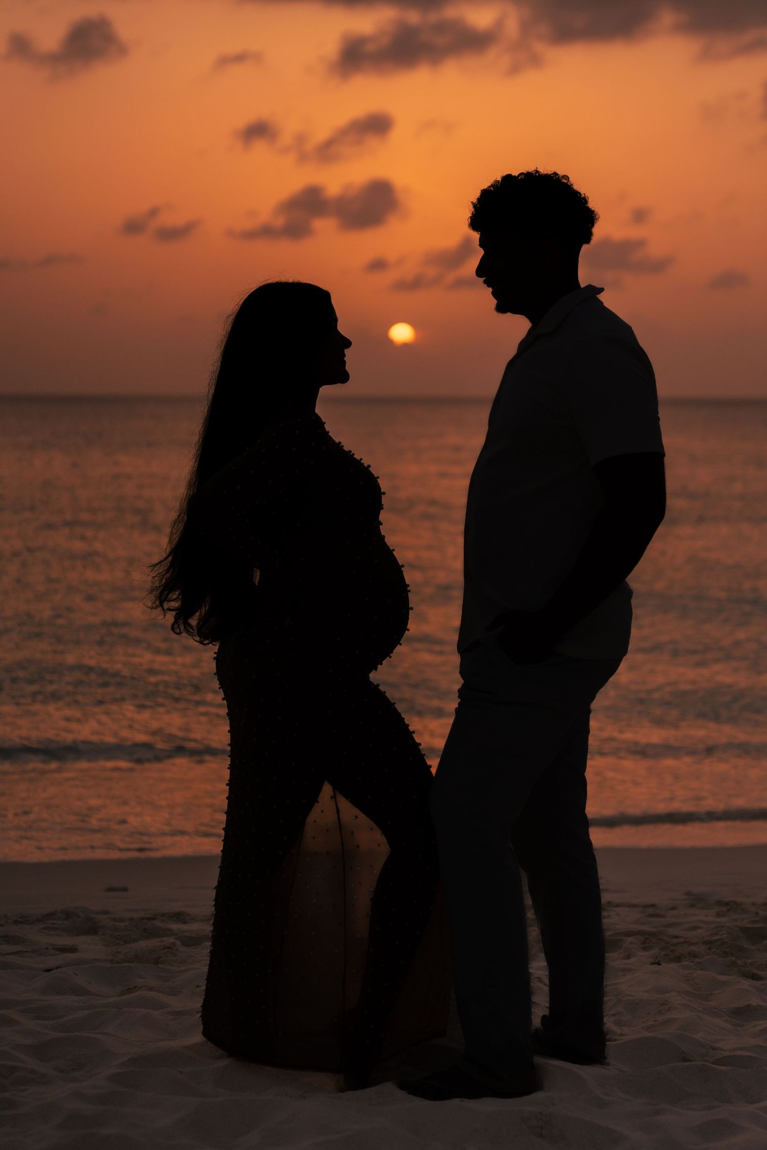 Silhouette of expecting couple standing on beach against dramatic orange sunset.
