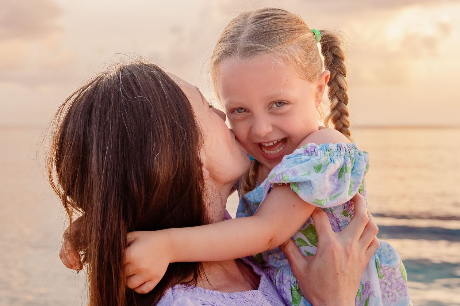 A mother lovingly kisses her laughing little girl with braids on a beach at sunset.