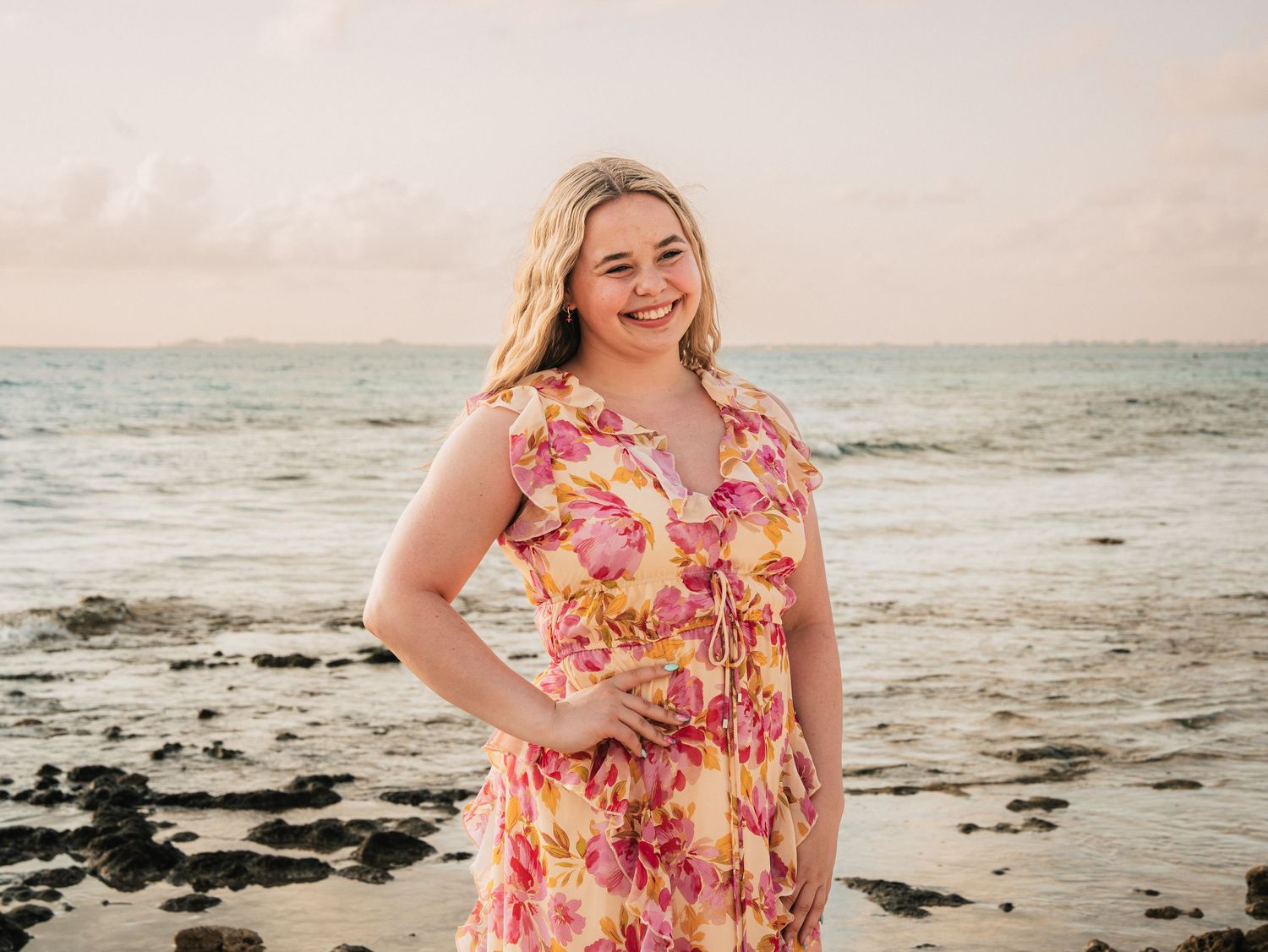 A young blonde woman in a yellow and pink floral dress poses confidently on a rocky beach at sunset.