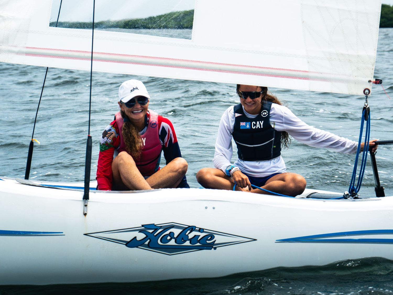 Two women sailing on a Hobie Cat sailboat on open water, wearing life vests and sunglasses.