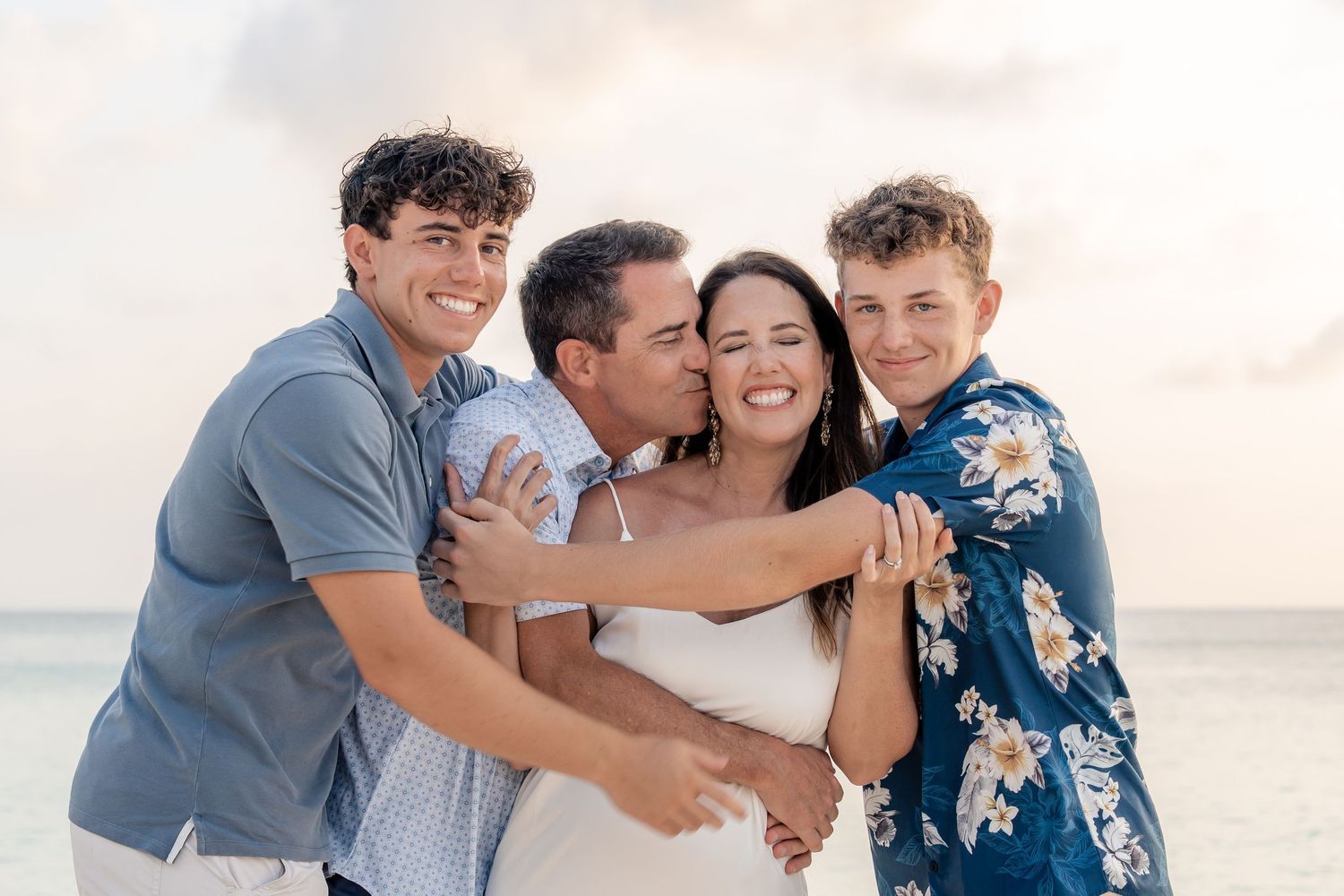 Family of four smiling and embracing on a beach at sunset, with ocean waves in the background.