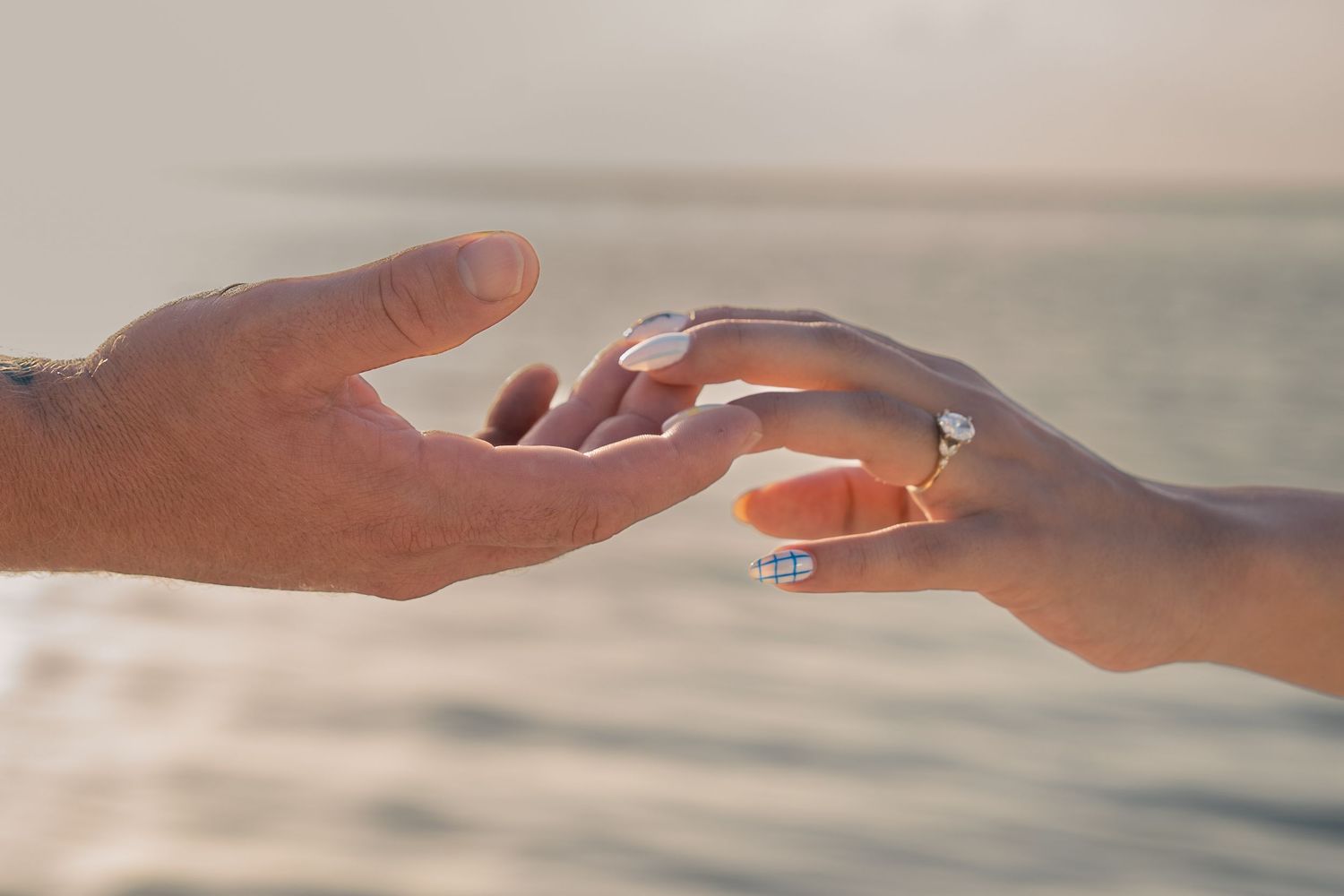 Two hands reaching toward each other near the ocean, one wearing a diamond engagement ring, symbolizing a romantic proposal.