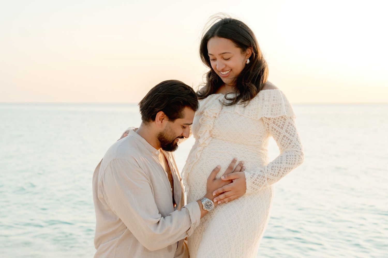Couple sharing a tender moment on the beach at sunset, with the woman in a white lace dress showing her baby bump.