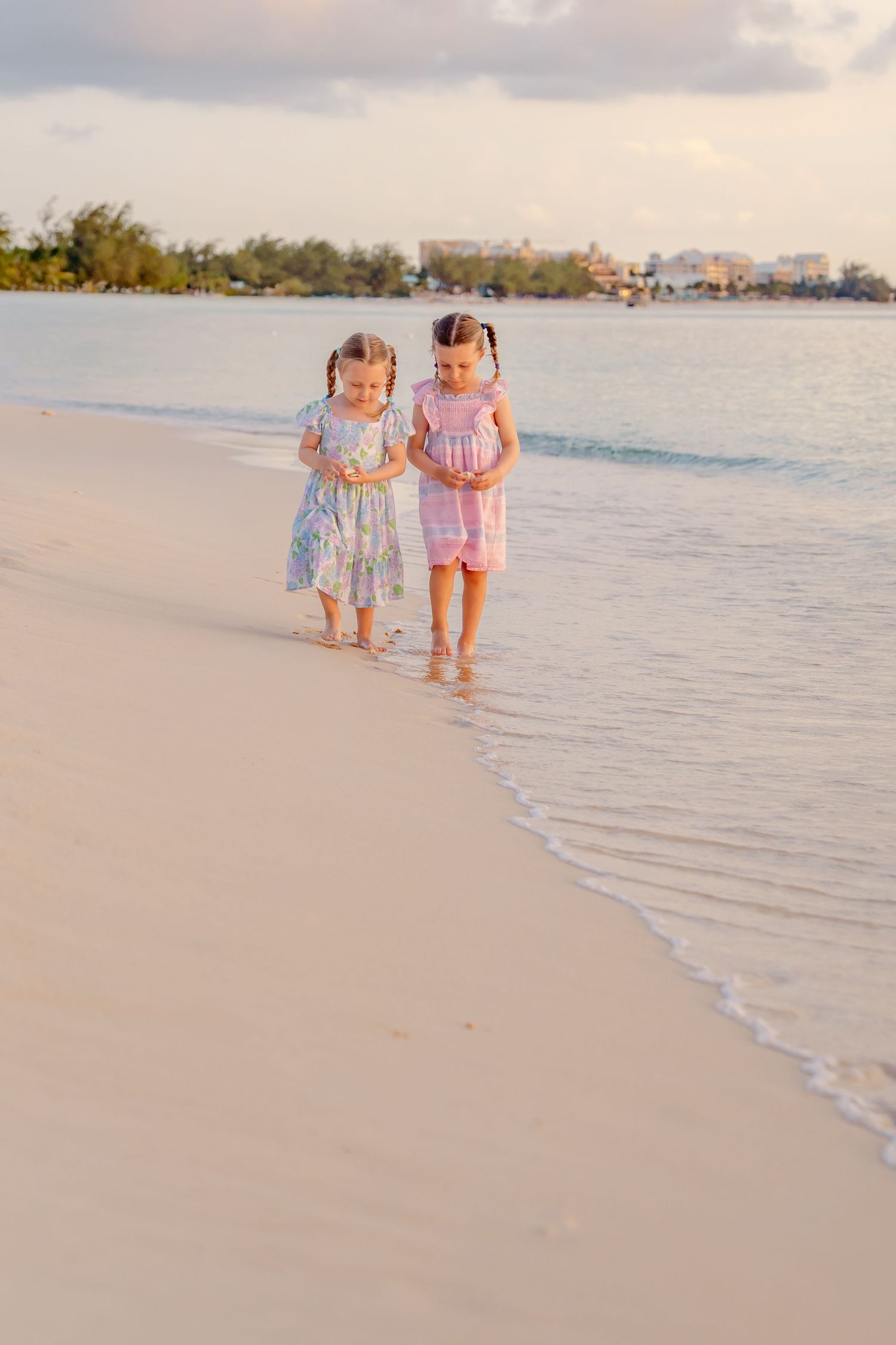 Two young girls in floral dresses walk along a sandy beach at sunset near calm ocean waves.