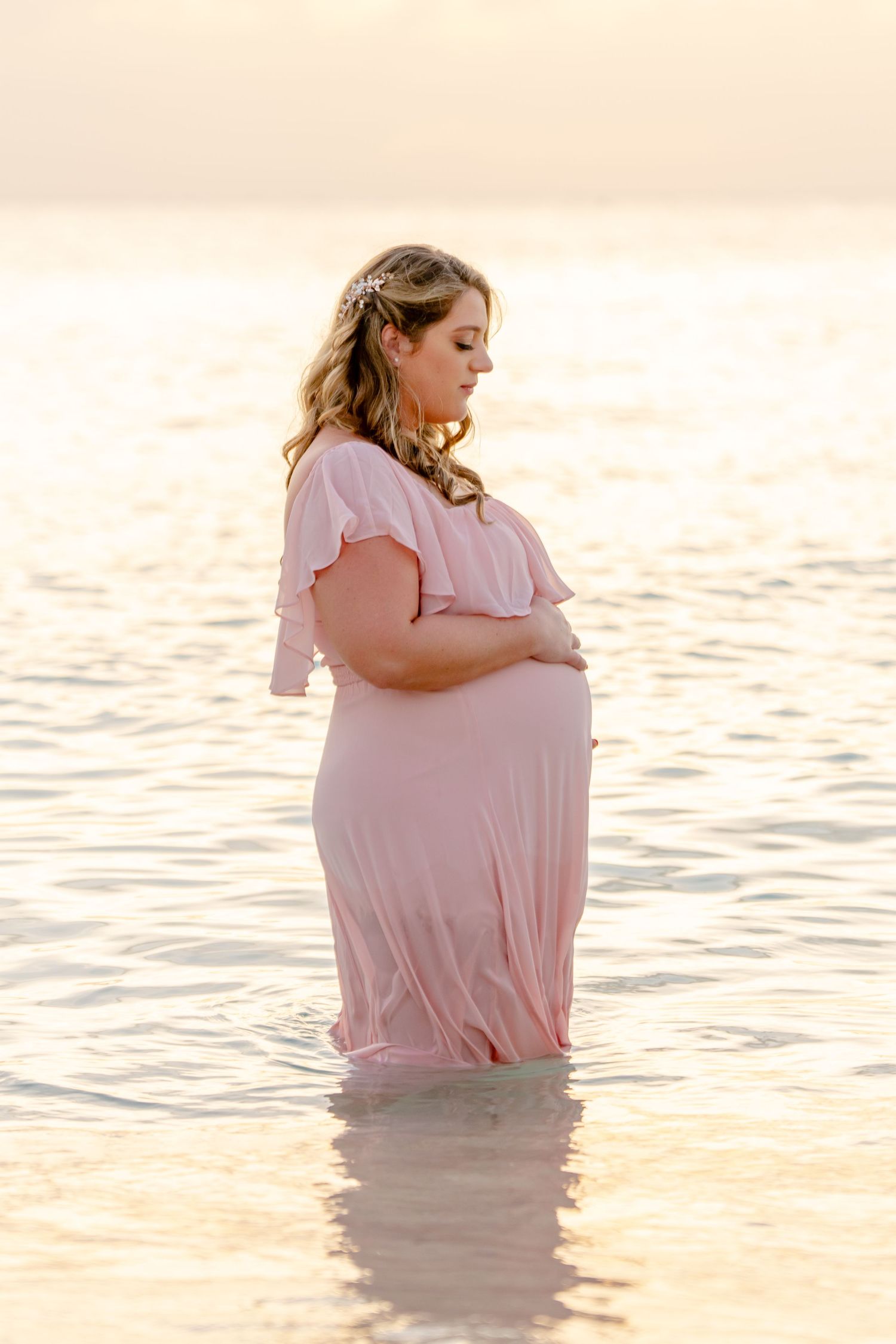 Person in pink dress stands in shallow ocean water during sunset maternity photoshoot.
