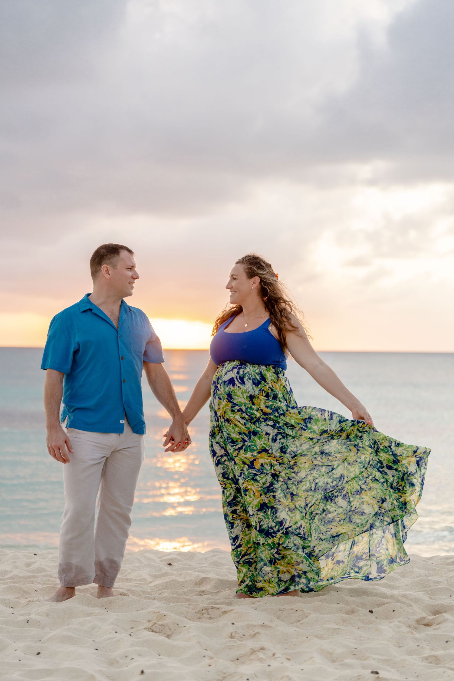 Romantic beach photoshoot with couple in blue and floral dress walking along shoreline at sunset.