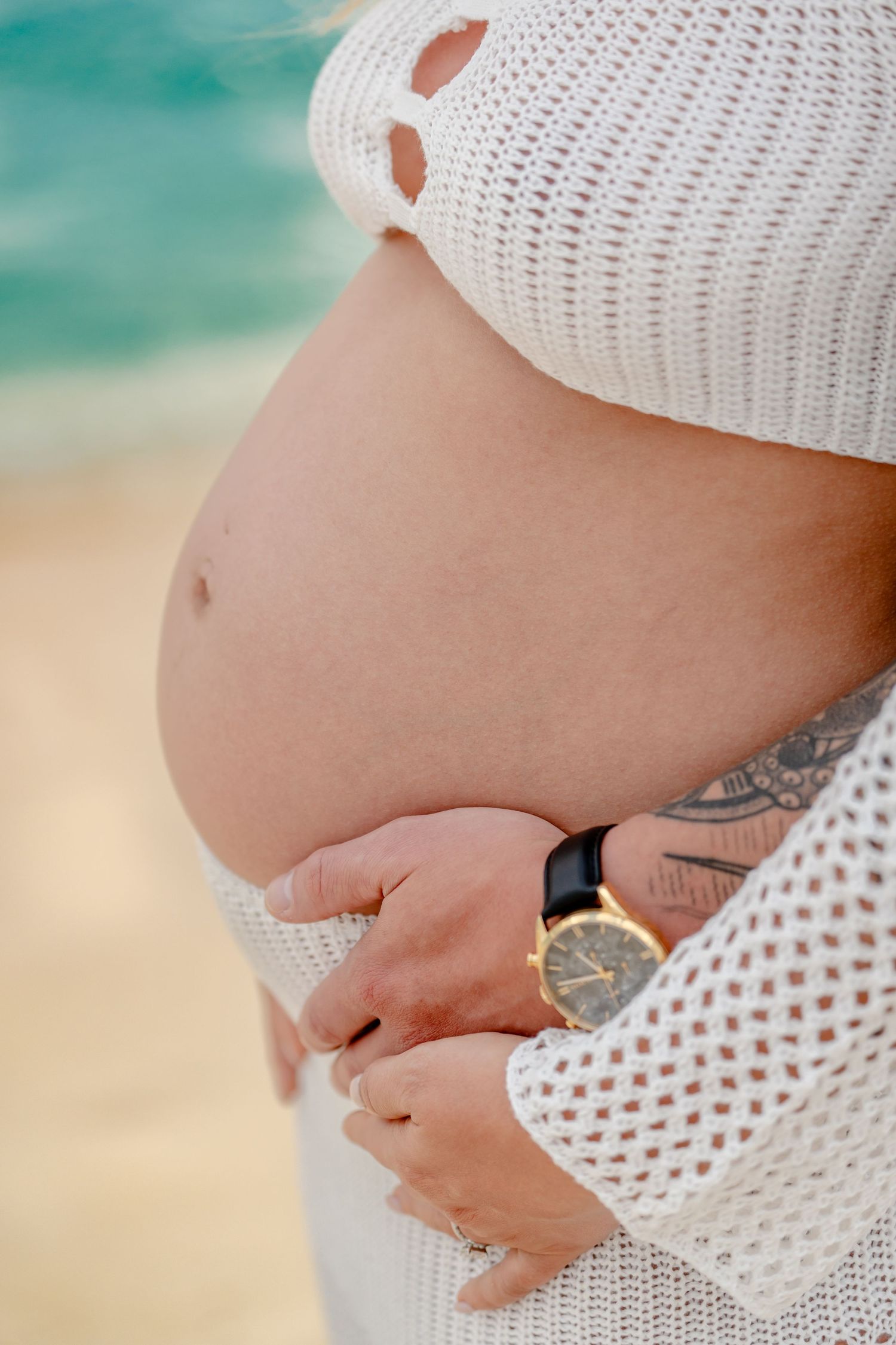 Close-up details of hands cradling pregnant belly in white knit dress by the ocean.