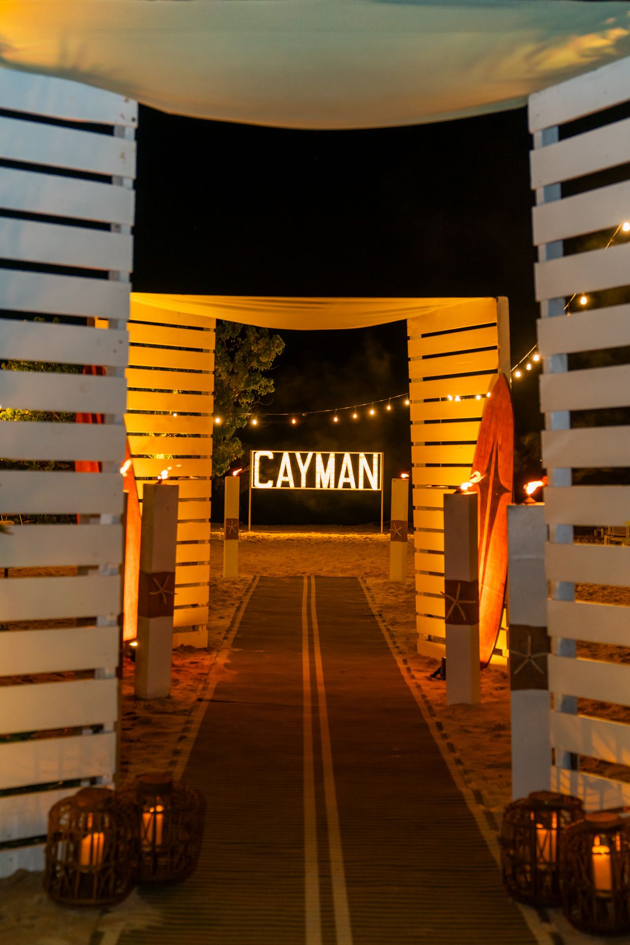 Illuminated wooden walkway with string lights and Cayman signage creating an atmospheric evening path.