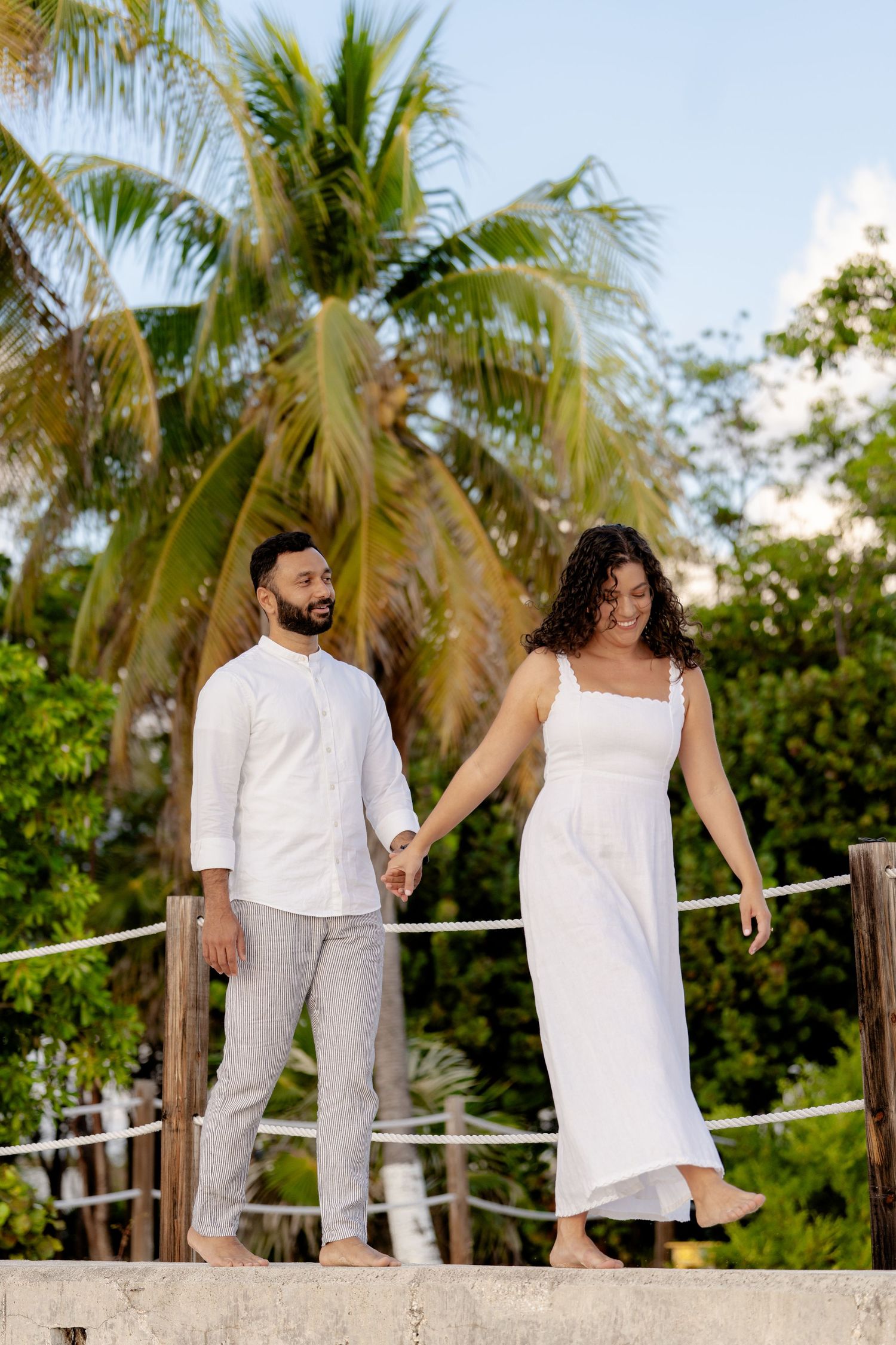 A couple in white outfits holding hands while walking on a tropical boardwalk near palm trees.