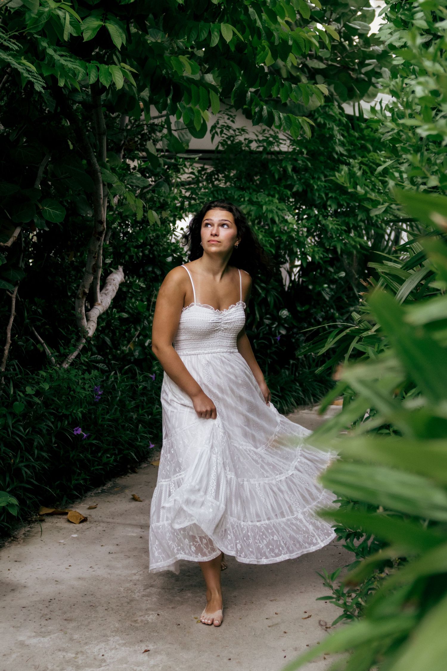 Someone twirls in a white lace dress among tropical greenery and garden paths.